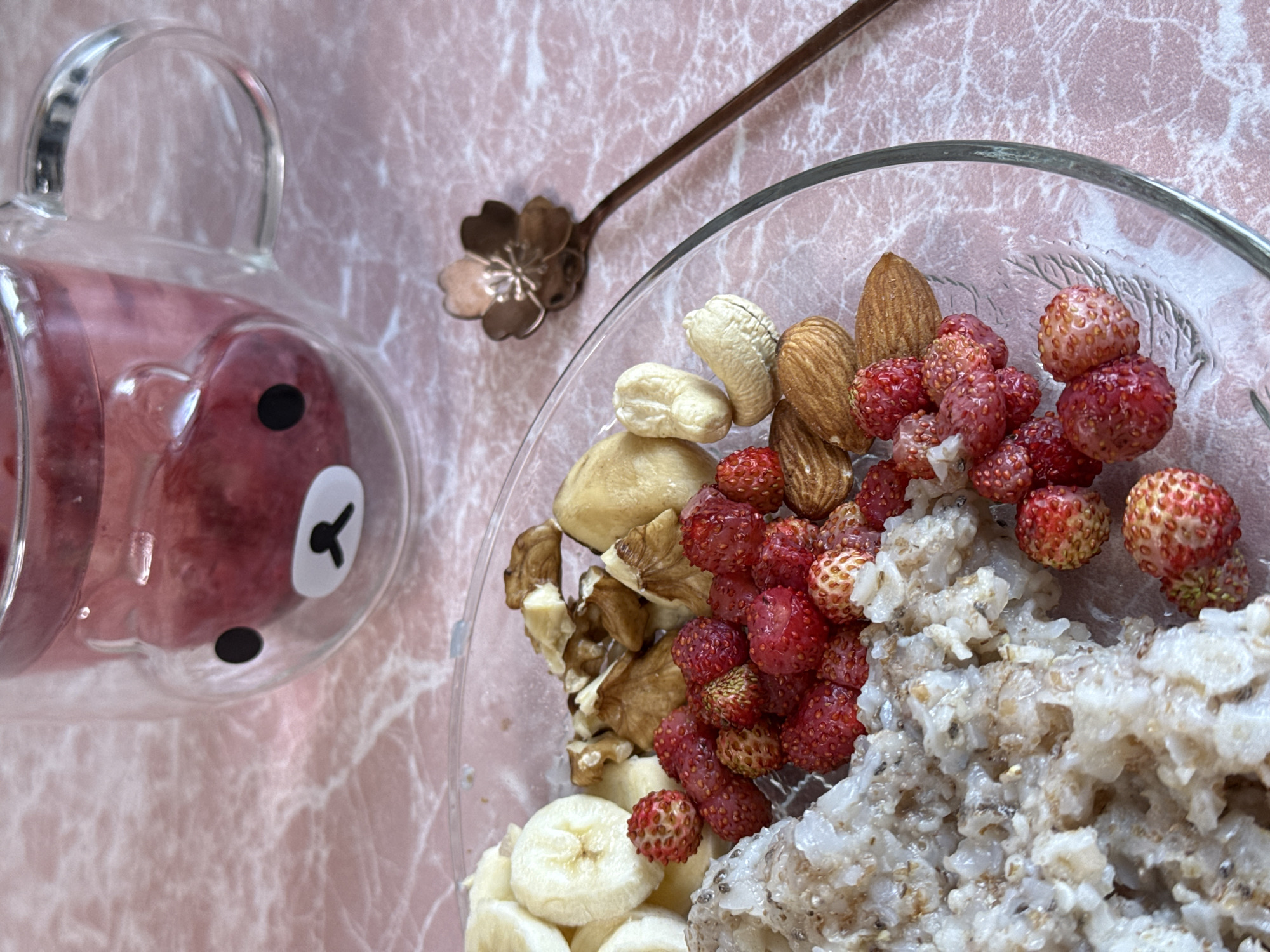 A clear glass mug with a double-walled design, featuring a caricature of a bear's face on the exterior, is filled with a pink liquid and sits on a pink marbled surface. Next to the mug, a clear glass bowl contains oatmeal, sliced bananas, walnuts, cashew nuts, almonds, and wild strawberries. A small, rose-gold colored spoon with a floral motif rests on the right side of the bowl, pointing towards the mug. The scene is shot from a slightly elevated angle.
