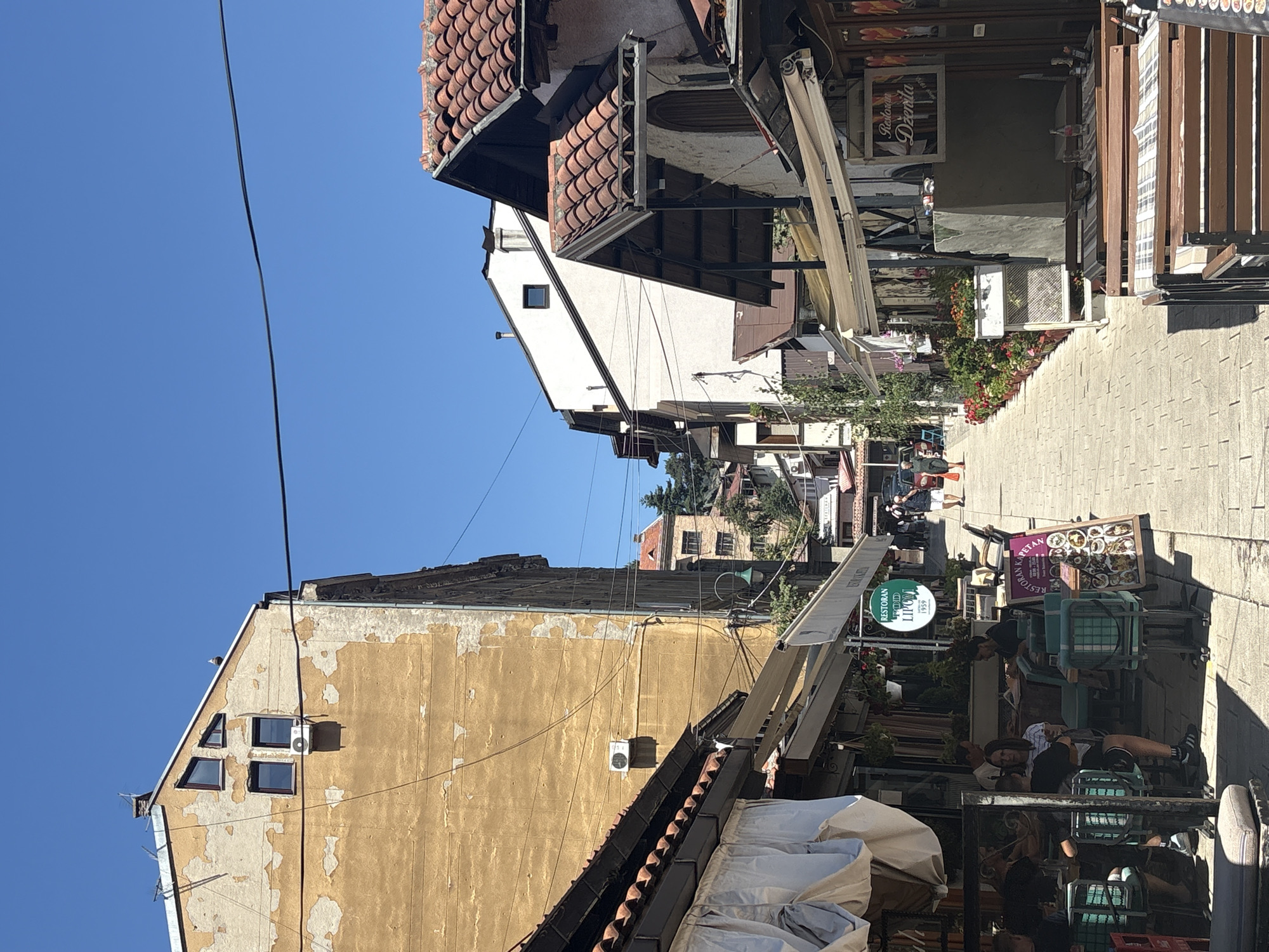 The image depicts a street scene in Sarajevo, Bosnia and Herzegovina, likely in an area with outdoor cafes and restaurants. The architecture suggests a blend of older and newer buildings. In the foreground, the left side shows the exterior of a restaurant with white awnings and outdoor seating. People are seated at tables. A green sign for "RESTORAN FOOD LIPONT 1959" is visible. The right side of the street also features outdoor dining setups with awnings and tables. A sign for "Restoran Džirlo 1970" is present. The street is paved with cobblestones and lined with potted plants. Several people are walking along the street in the background. The buildings vary in height and architectural style, with some showing signs of age. The sky is clear and blue, indicating a sunny day. The overall impression is that of a lively urban area during daytime.