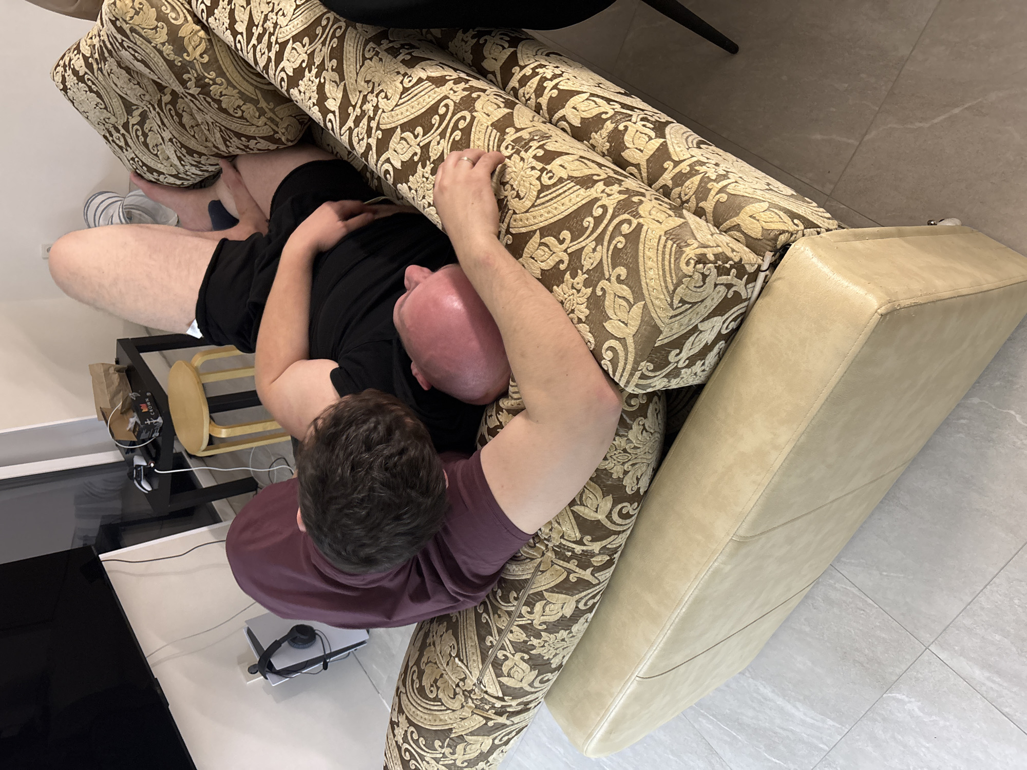A man lies face down on an ornately patterned couch. He is wearing a maroon shirt and black shorts. The couch is positioned in a room with tiled flooring, near a white table that holds a black television and a PlayStation 5. The location is presumed to be an interior setting.