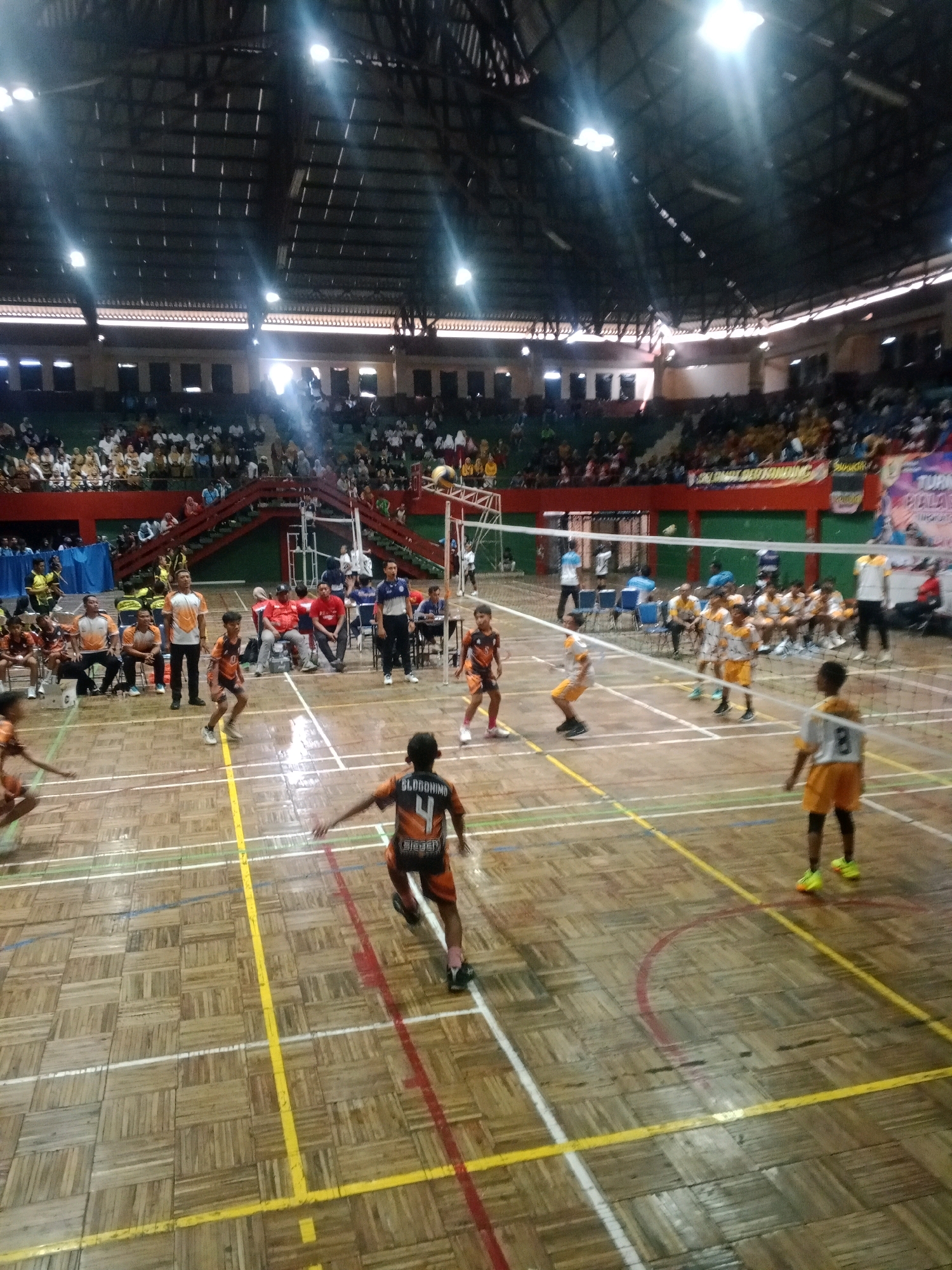 An indoor volleyball game is underway in a gymnasium in Wuryorejo, Indonesia. The court is made of wood with yellow and red lines marking the boundaries and areas. A net divides the court in half. Spectators are seated in tiered seating surrounding the court. Several young players are actively engaged in the game, some in orange and black uniforms and others in yellow and white. One player in the foreground is wearing an orange jersey with the number 4 and the name "SLOGOONIMO" printed on the back, and is in the motion of playing. Another player in the background is wearing a yellow jersey with the number 8. The lighting in the gymnasium is bright, with overhead lights illuminating the court. The overall scene depicts a competitive sporting event with a focus on youth participation.