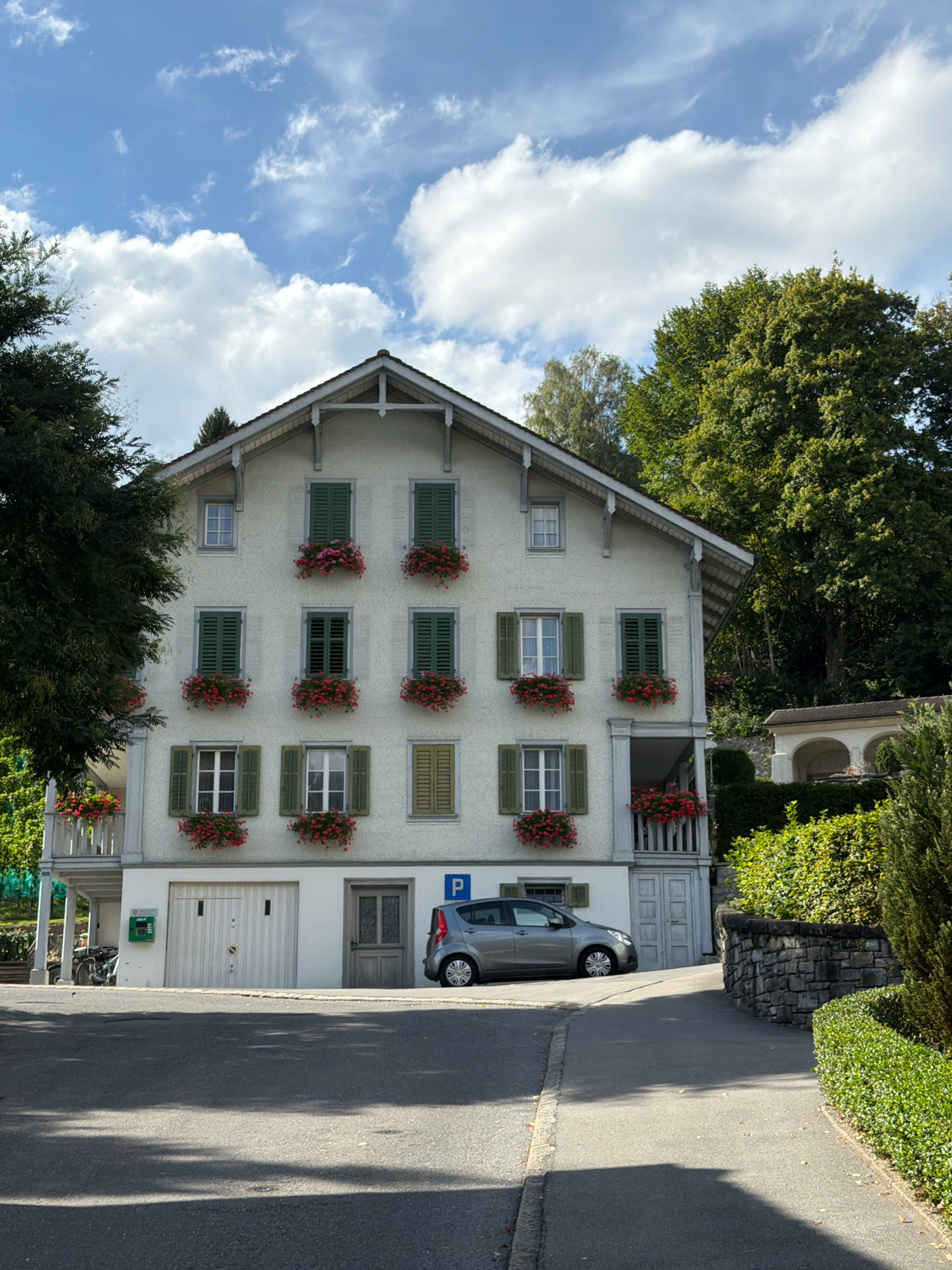 A three-story, light-colored building with a peaked roof is the main subject of the image. The building features numerous windows with green shutters and flower boxes filled with red flowers. On the ground floor, there is a large garage door and a smaller side door. A silver sedan is parked to the right of the garage door, and a bicycle is parked to the left. The building is surrounded by trees and lush greenery. The sky is blue with scattered white clouds. The scene appears to be a residential area with a paved road in the foreground.