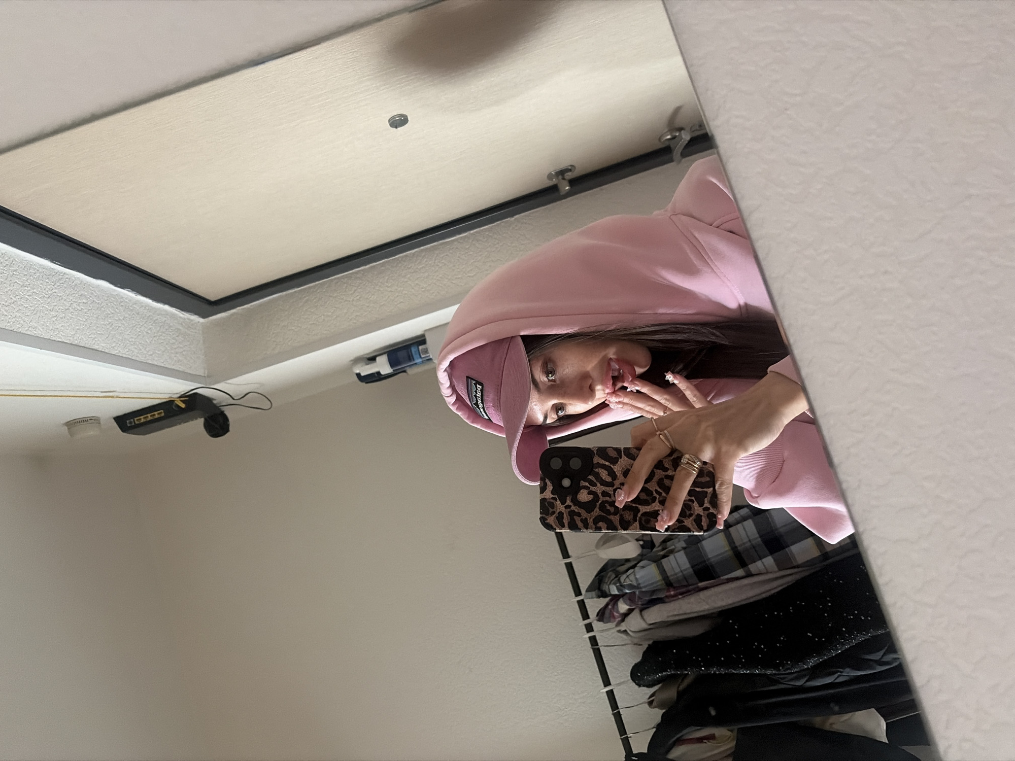 A young woman, reflected in a mirror, poses for a selfie in what appears to be an indoor domestic setting, possibly an apartment hallway in Saint Petersburg, Russia. She is wearing a pink hooded sweatshirt and a pink baseball cap. Her left hand, adorned with rings and long nails, holds a smartphone with a leopard print case, angled to capture her reflection. Her mouth is slightly open, with her tongue visible, and she appears to be sticking her tongue out or touching her lip with it. In the background, visible in the mirror, is a rack of clothing, suggesting a closet or wardrobe area. To the right of the woman, a doorframe and part of a door are visible, consistent with an interior doorway. Above the rack of clothes, a black network switch is mounted on the wall. The overall impression is of a casual, personal moment captured by the woman herself.