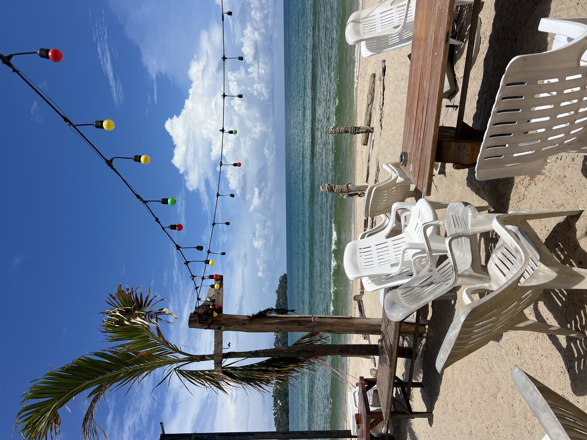 A bright, sunny day on a sandy beach with a calm ocean and blue sky dotted with white clouds. String lights with colorful bulbs hang overhead. In the foreground, several white plastic beach chairs surround wooden tables. A palm tree stands to the left, with a wooden post and some fishing net nearby. In the background, the ocean stretches to the horizon, with a distant green, tree-covered landmass visible on the left.