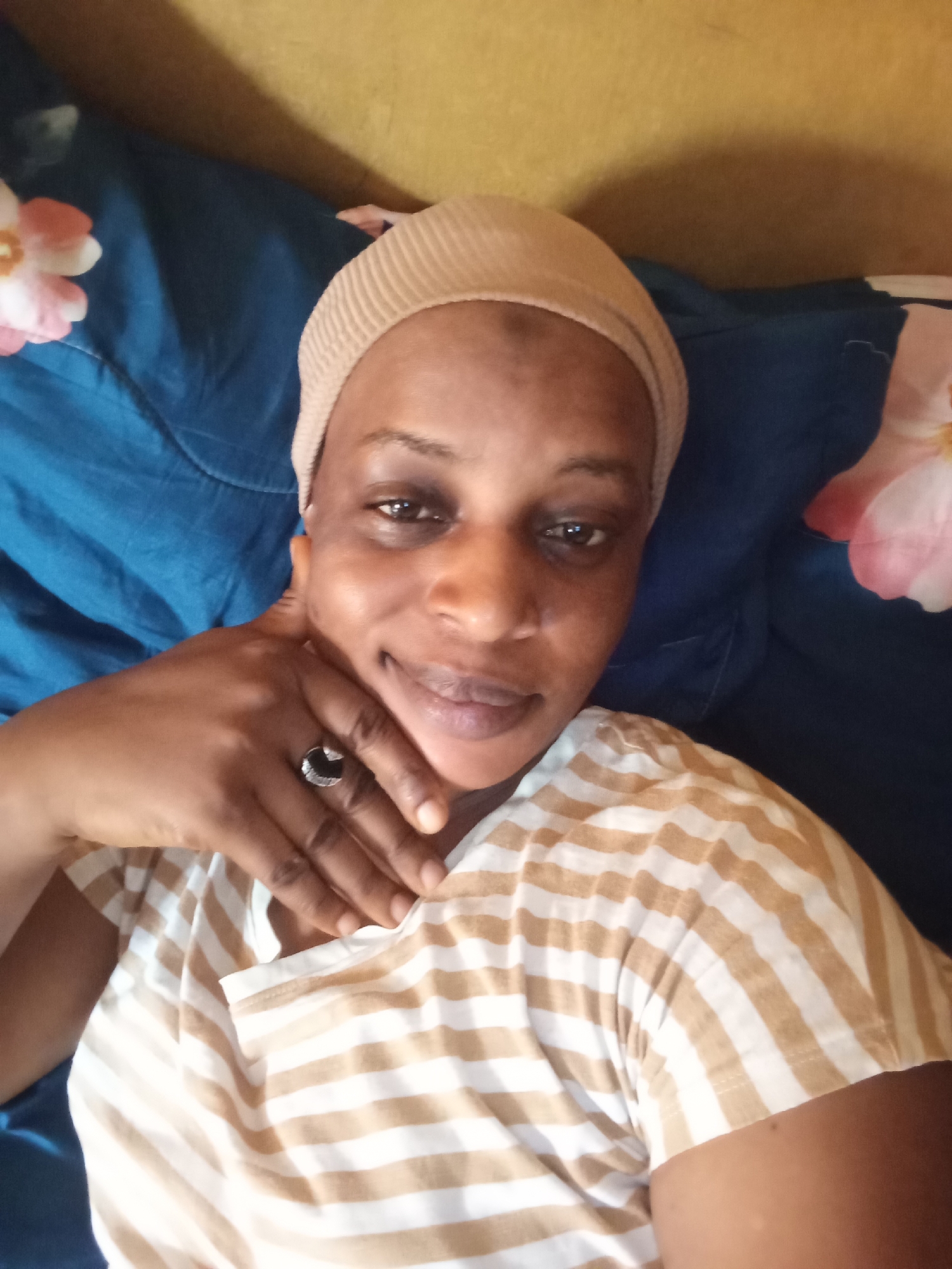 A medium shot features an African woman lying in what appears to be a bed. She is wearing a beige head covering and a striped t-shirt with tan and white stripes. Her left hand is raised to her chin, and she is wearing a ring with a dark stone on that hand. She is looking directly at the camera with a slight smile.
The bed has a dark blue comforter with light pink flowers. The background appears to be a solid yellow wall.
The information about the location in the city of Narayi, Nigeria, is not visible in the image.