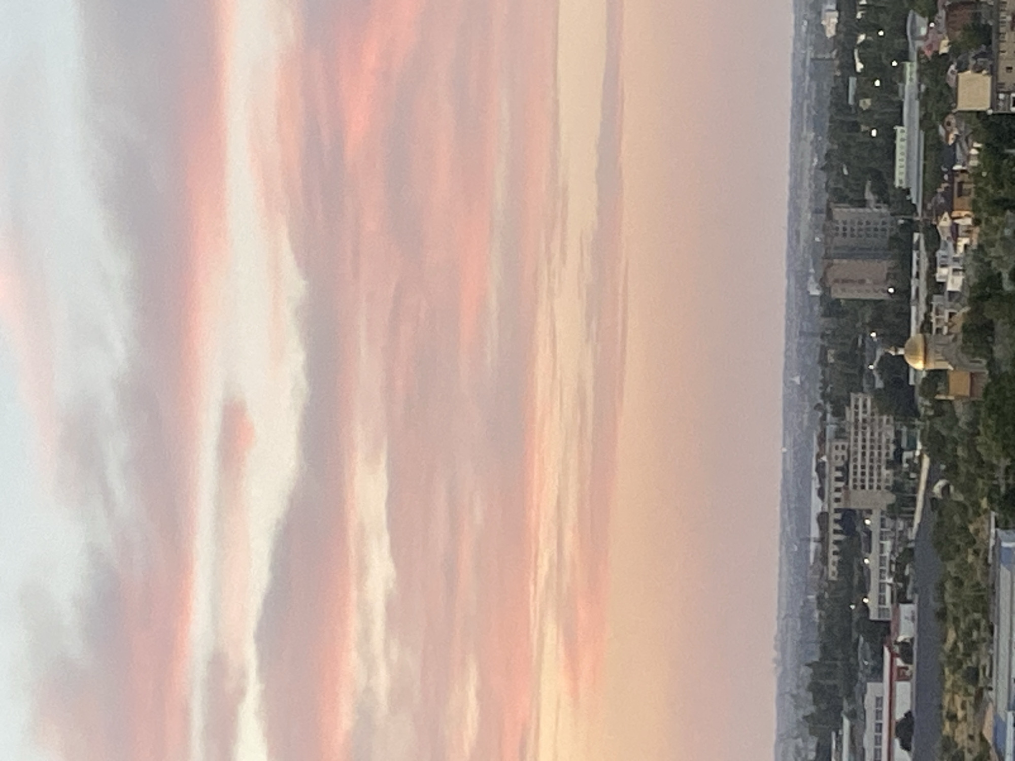 This is an aerial, eye-level, medium shot of a city in Chornomorsk, Ukraine, at dusk. The sky is filled with soft, pinkish-orange clouds. Below the sky, a cityscape is visible with buildings, trees, and the faint outlines of distant structures. The overall atmosphere is calm and serene.