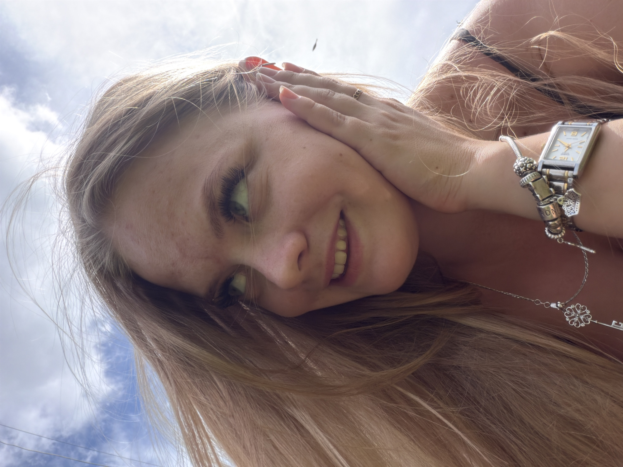 The image is a close-up, eye-level shot of a young Caucasian woman outdoors. She has long, blonde hair that is being blown by the wind, framing her face. Her left hand is touching her cheek, and she is looking to her left. She is wearing a silver watch with charms on her left wrist, and a necklace with a key pendant is visible on her chest. The background is a sky with clouds and visible overhead wires, suggesting an urban or semi-urban outdoor setting. The lighting indicates it is daytime, possibly sunny. No specific landmarks identifiable to Chaikovsky, Russia, are present in the image.