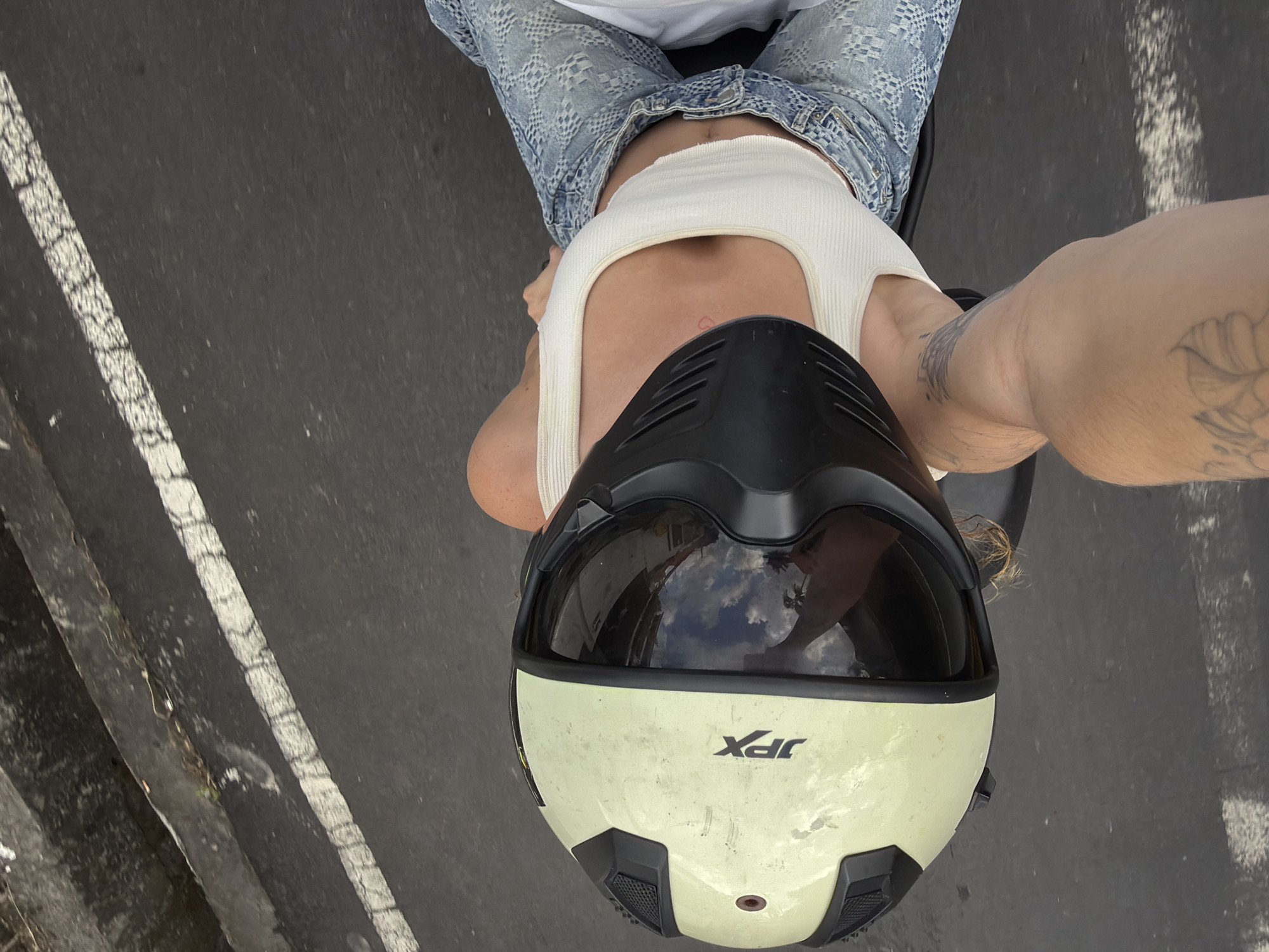 An overhead, full shot captures a person wearing a light yellow and black full-face motorcycle helmet while seated on a motorcycle. The person is dressed in a white tank top and distressed denim jeans. Their right arm is visible, with tattoos on the forearm. The helmet's visor reflects the sky and palm trees, indicating an outdoor setting, possibly in a tropical environment. The motorcycle is on a paved road with white lane markings. The background shows a concrete barrier or ledge. The scene appears to be in North Kuta, Indonesia, based on the context provided.