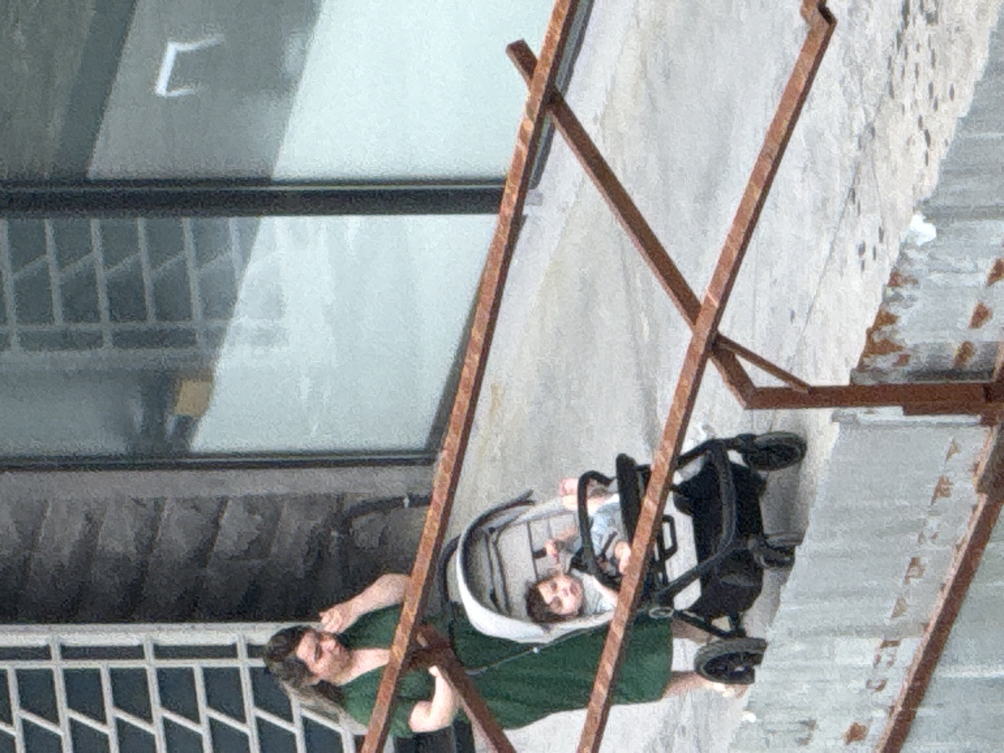 A person in a green shirt is pushing a stroller with a baby inside. The stroller is grey and black. The person is on the left side of the image and is looking to the right. The baby is in the stroller, lying on its back. The background is a concrete wall and some metal bars. The scene appears to be outdoors during the day. There is no visible text in the image. The overall mood appears casual and everyday.