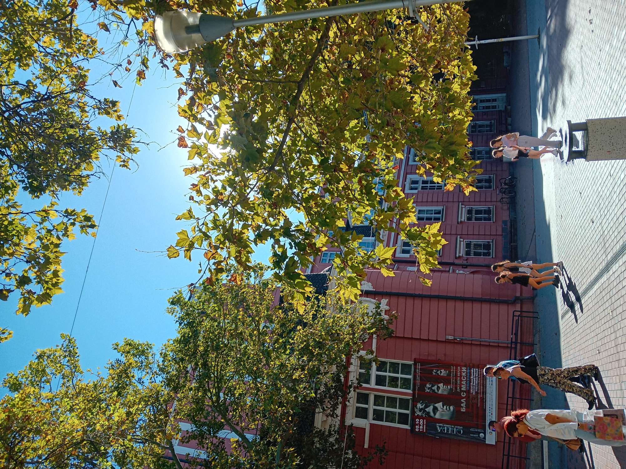 This media file depicts an outdoor scene in Varna, Bulgaria, bathed in bright sunlight, suggesting a clear day. The image is oriented vertically, with trees and a building dominating the foreground and midground. The building is a striking red, featuring multiple windows with white frames, and appears to be a multi-story structure. A poster is visible on the building's facade, advertising an opera performance of "Un ballo in maschera" by Giuseppe Verdi, with a date and time indicated as "02.10 19:00".
Several individuals are present on a paved walkway in front of the building. Three people are walking away from the camera, and a group of three are walking towards the camera. The people in the foreground appear to be engaged in conversation, with one person looking towards the camera. The dappled sunlight filtering through the trees creates a dynamic play of light and shadow on the ground. The presence of trees with autumn foliage suggests the image may have been taken in the fall. The overall atmosphere appears relaxed and leisurely, characteristic of a daytime stroll in a city environment. A lamppost is also visible, adding to the urban setting.