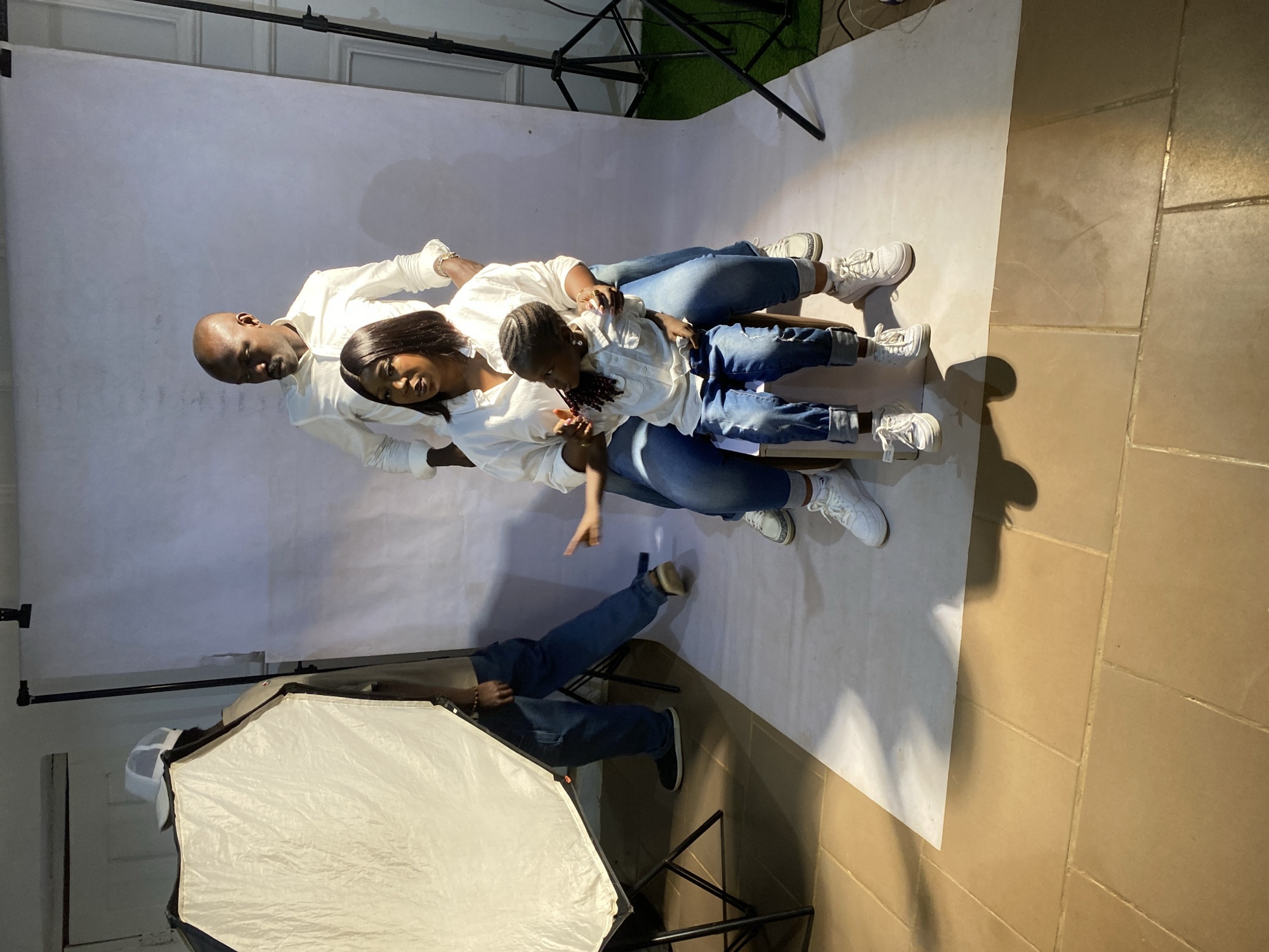 A family of three is lying on a white backdrop. The father is on the left, followed by the mother and then the child. All three are wearing white shirts and blue jeans. The father is bald. The mother has straight, dark hair. The child has braided hair. A photography light and other photography equipment are visible in the frame. The floor appears to be tiled. The photo was taken inside a room or studio, in the city of Jos, Nigeria. There is another person in the background.