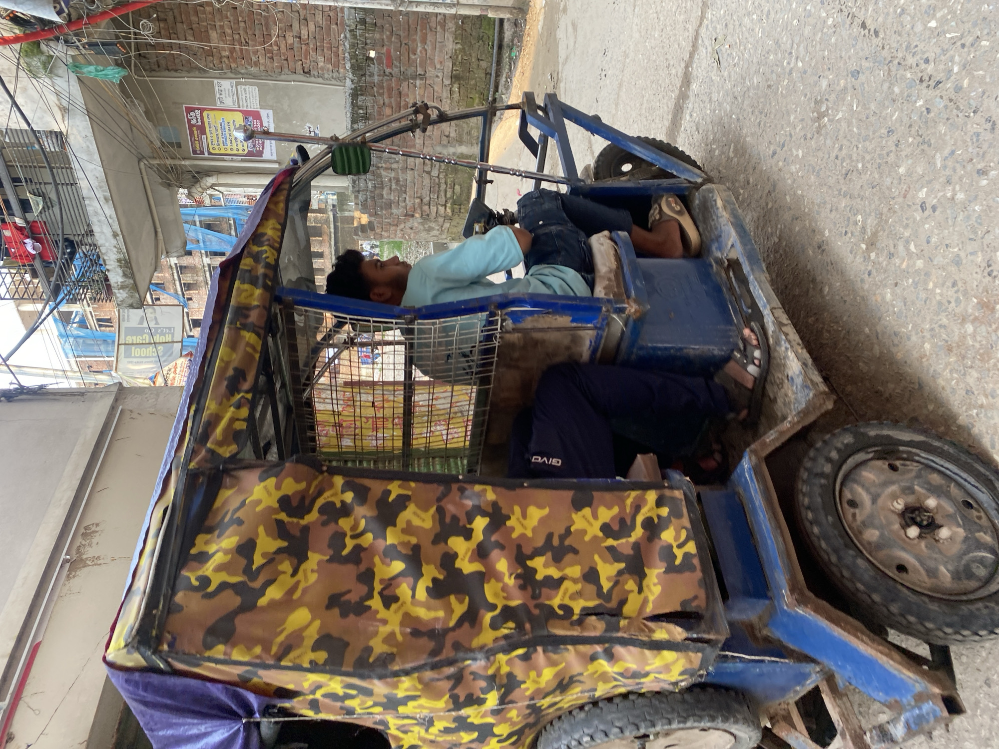 Here's a description of the image:
The image depicts a man reclining inside a blue, makeshift auto-rickshaw. The rickshaw has a camouflage-patterned cover and is parked on a street. Another person is seated in the back part of the rickshaw. The background shows a building with a brick wall and an advertisement in Bangla. Wires run along the buildings. It appears to be a street scene in Dhaka, Bangladesh.