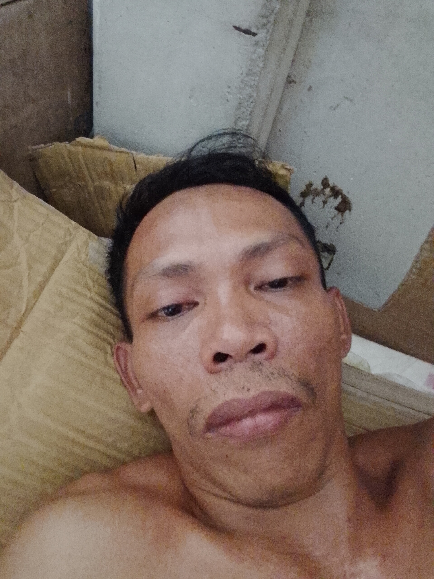 A medium close-up shot of a man lying down, looking directly at the camera. The man is likely in his 30s to 40s, with dark hair, dark eyes, and stubble on his upper lip. His complexion appears weathered. He is shirtless. The background consists of cardboard and a concrete surface, potentially indicating a makeshift bed or sleeping area. No specific details in the image confirm the location as Bukit Ketok, Indonesia.