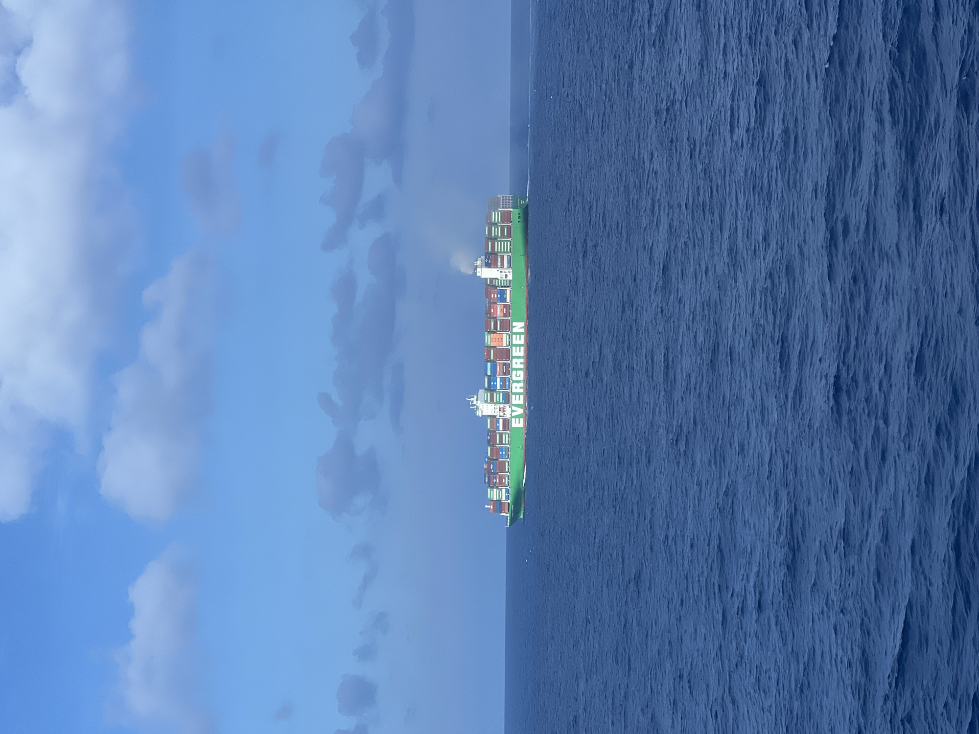 A large, green Evergreen container ship is sailing on a dark blue ocean under a blue sky with scattered white clouds. The ship is filled with numerous stacked shipping containers of various colors, including red and white. Smoke is emanating from the ship's smokestack. The water is choppy with visible waves. The image appears to be taken from a distance, likely from another vessel or a high vantage point. The location is unknown, and no people are visible.