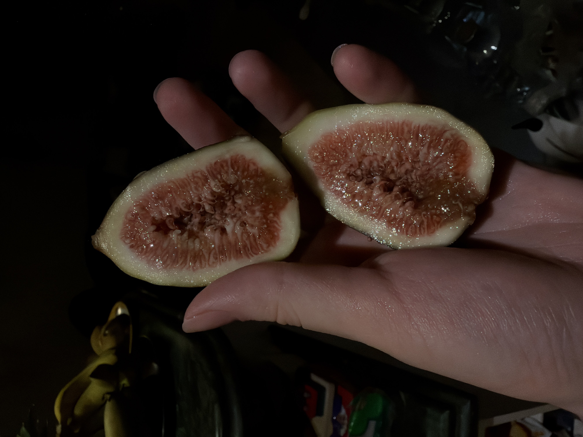 A close-up shot shows a person's hand holding two halves of a ripe fig. The figs are cut lengthwise, revealing a vibrant pink, juicy interior with numerous small seeds. The skin of the figs is a pale green to yellowish color. The hand is positioned to display the cut surfaces of the figs, with fingers gently supporting them. The background is dark and out of focus, with some blurry objects visible, including what appears to be a bunch of bananas on the left and some indistinct items on the right. The lighting is dim, highlighting the textures and colors of the figs and the skin of the hand. There is no indication of the city of Sharm El Sheikh, Egypt, in the image.