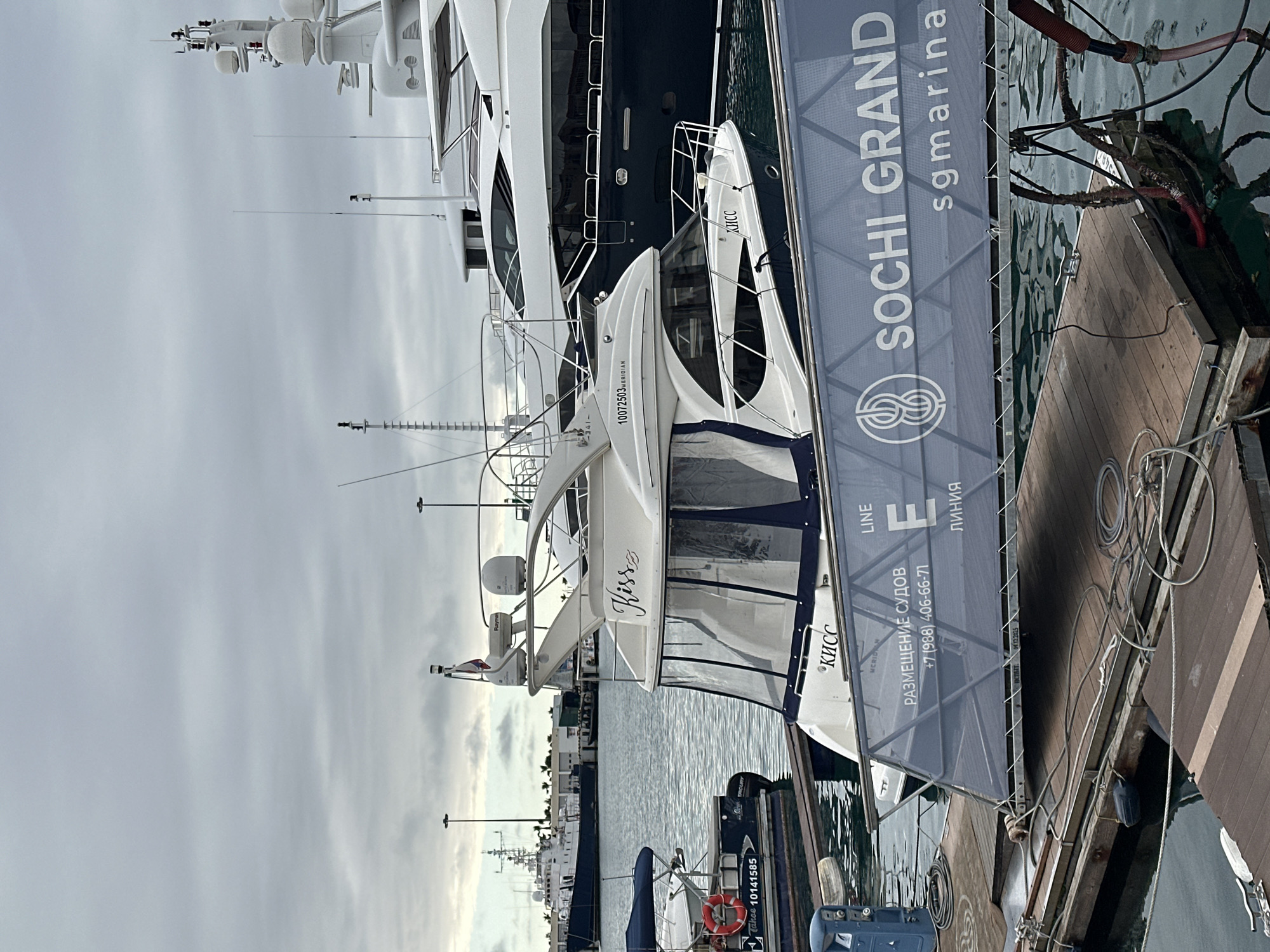 The image depicts a marina scene under an overcast sky in Sochi, Russia, specifically at the Sochi Grand Marina. In the foreground, a white motor yacht named "Kiss" (also bearing "КИСС" in Cyrillic) with registration "10072503 MERIDIAN" is moored to a wooden floating dock. This yacht features a clear vinyl enclosure, an antennae mast with "Raymarine" equipment, and a small Russian flag. A blue banner, acting as a gangway, partially obstructs the view of the dock and the yacht's hull, displaying "SOCHI GRAND sgmarina," "LINE E" ("ЛИНИЯ"), and contact information for "РАЗМЕЩЕНИЕ СУДОВ" (Vessel Accommodation) at "+7 (988) 406-66-71".
Behind the "Kiss" yacht, a much larger, dark blue-hulled superyacht is also docked. To the left, a smaller, dark blue pontoon-style boat named "TAKEE 10141585" with a red lifebuoy is moored. Further in the background, across the water, a grey ship, possibly a patrol or utility vessel, is visible near distant port infrastructure and buildings. The wooden docks show coiled ropes, mooring cleats, a blue utility pedestal, and various hoses. The water is calm, reflecting the cloudy sky. No individuals are visible in the scene.