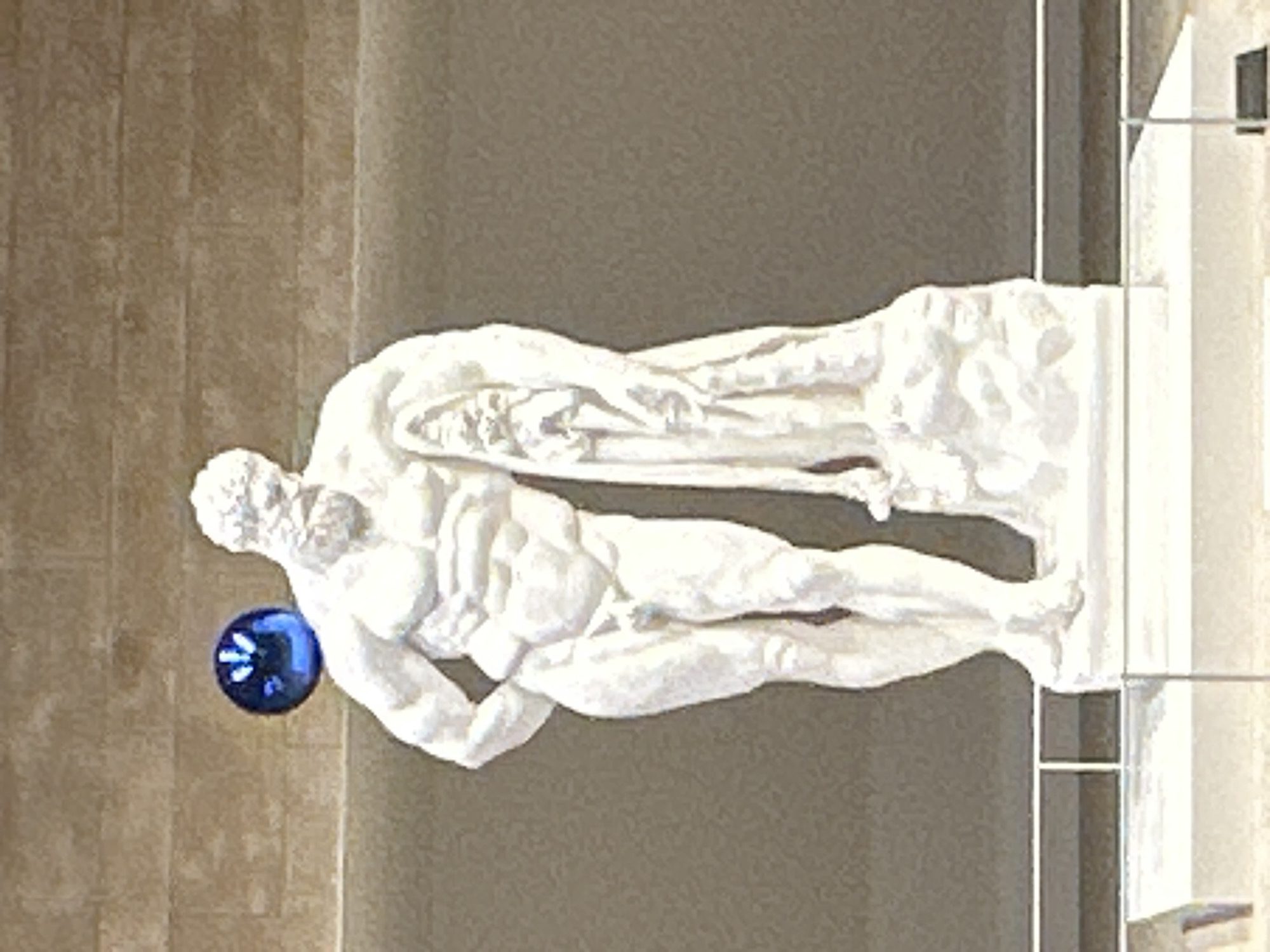 A white marble sculpture of a muscular man is displayed on a white pedestal. The man is leaning to his right, with his right hand on his hip and his left arm draped over a piece of fabric that falls to his side. He has a beard and is depicted with idealized musculature. Behind the sculpture, a blue, reflective spherical object is suspended in the air, partially obscuring the sculpture's shoulder. The background appears to be a wall with horizontal, textured panels in a light brown or tan color. The lighting is even, suggesting an indoor setting such as a museum or gallery. There is no identifiable location in the city of Incheon, South Korea, associated with this image.