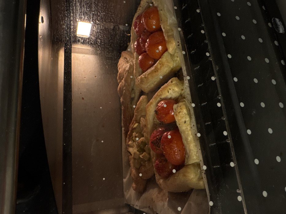 The image displays the interior of an operational oven, illuminated by an internal light source positioned on the upper right. A metal baking tray, lined with off-white parchment paper, occupies the central focus on an oven rack. On the tray, multiple pastry items are visible. In the foreground, two roughly oval-shaped pastries feature whole red tomatoes, some appearing softened and likely seasoned with dark specks. Behind these, a third pastry displays diced green fruit, consistent with apples. A larger, elongated pastry, possibly braided, rests further back on the tray. The lower portion of the oven's exterior, partially visible, is black with white polka dots. No individuals are present. The precise location within Tel-Aviv, Israel, cannot be determined from the visual evidence.