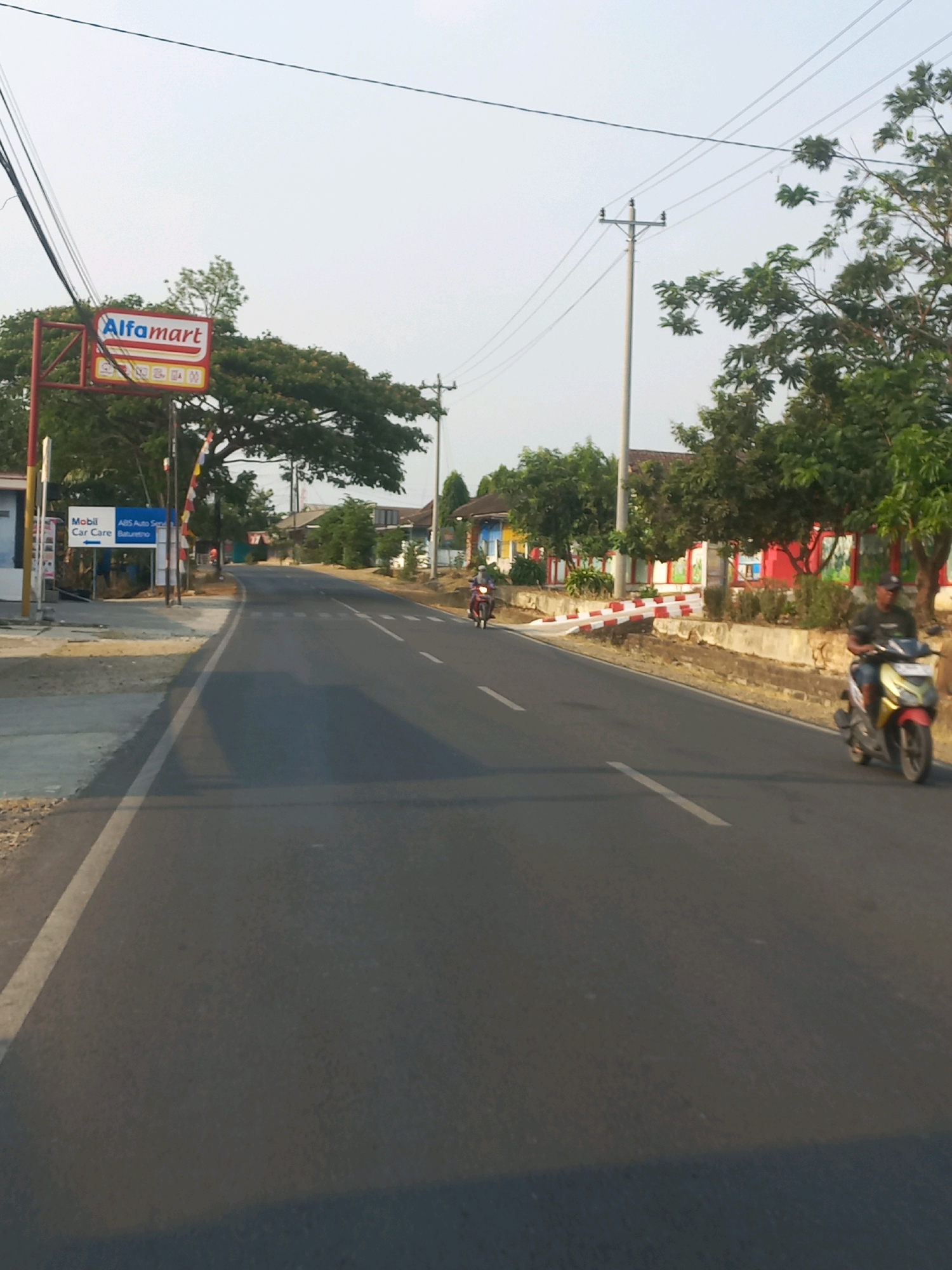 The image depicts a roadside scene in Baturetno, Indonesia, characterized by a paved road, commercial establishments, and residential buildings. On the left side of the road, an Alfamart convenience store is visible, identified by its prominent red and white signage. Adjacent to the Alfamart, a sign for "Mobil Car Care" with the text "ABS Auto Servis Baturetno" suggests an automotive service facility. The road itself is asphalted with white lane markings.
Several individuals are present, all operating motorcycles. One motorcycle rider, wearing a dark shirt and shorts, is moving towards the viewer on the right side of the road. Further down the road, another motorcycle is heading away from the viewer. The environment features numerous trees, some with dense foliage, lining the roadside. A utility pole with overhead wires stands on the right side of the road. In the background, buildings with colorful facades and red trim are visible, interspersed with more greenery. The sky is overcast with a light hue, indicating daytime. The overall impression is of a typical commercial and residential area in a town or city in Indonesia.