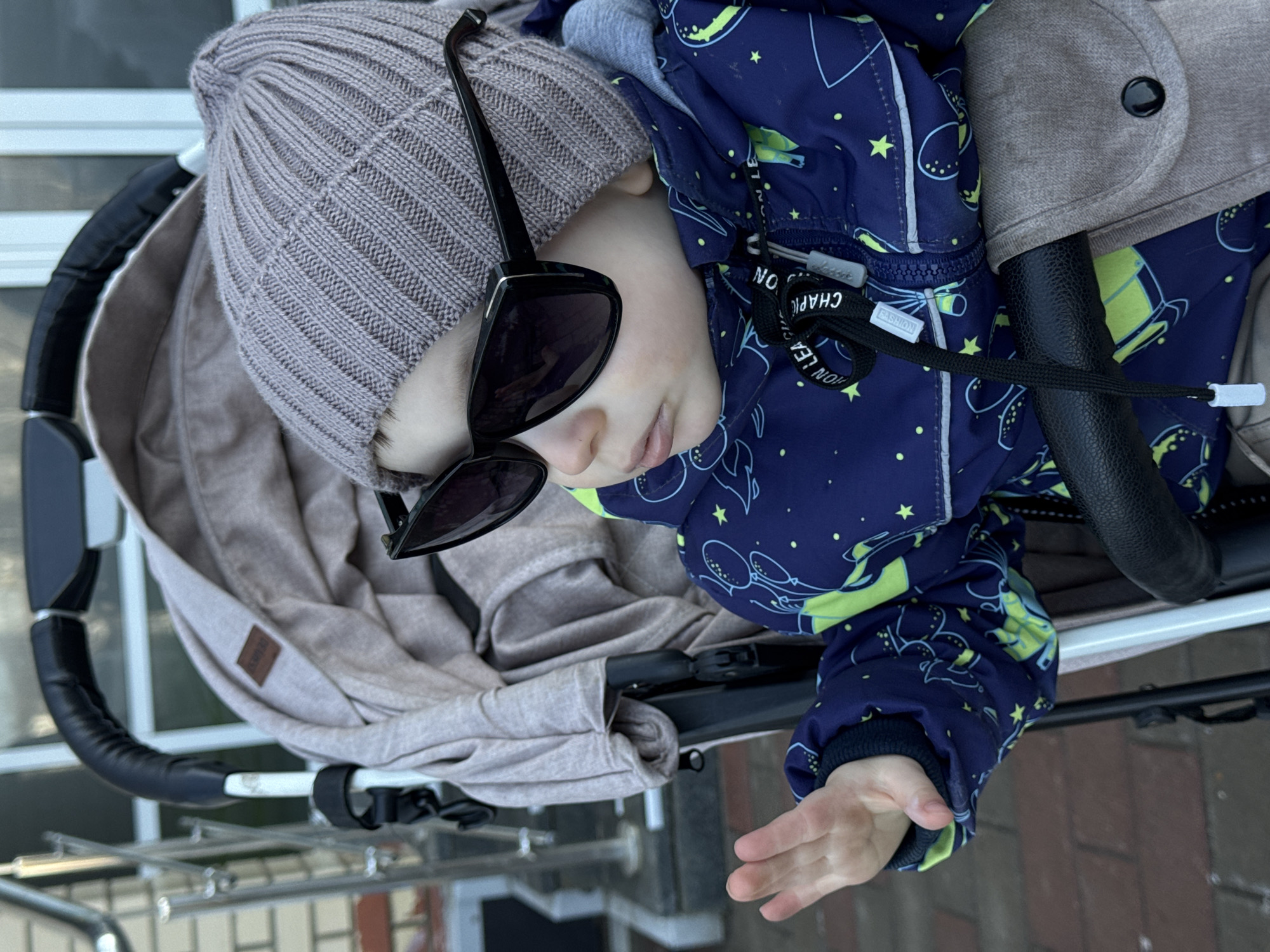 A young child lies in a stroller. The child is wearing a light grey knit hat and sunglasses. They also have on a blue jacket with a pattern of stars and planets. The stroller has a light brown interior lining and a black handlebar. The background suggests an outdoor location, possibly a street or walkway, with brick and metal elements visible. Based on the information provided, the location is Izhevsk, Russia.