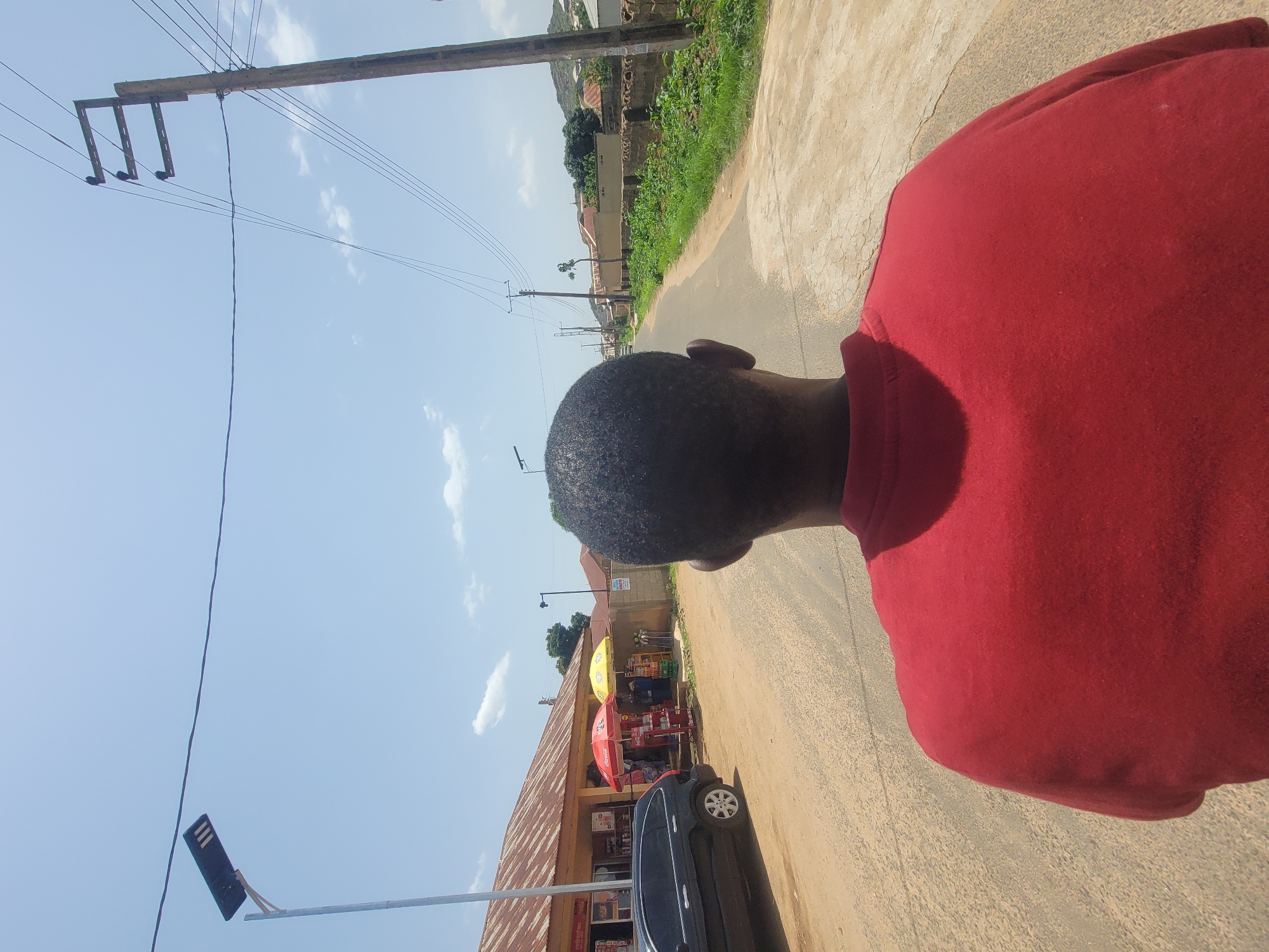 A person in a red shirt walks along a dirt road in Jos, Nigeria, on a sunny day. The person's head and shoulder are visible in the foreground, facing away from the camera. The road is lined with small shops on the left, some with red and yellow umbrellas providing shade. A dark car is parked on the side of the road. Electrical poles and wires are visible along the road, indicating power infrastructure. The sky is clear blue with a few wispy clouds. The overall scene suggests a typical day in a Nigerian town, with local commerce and daily life unfolding. The time of day appears to be daytime, given the bright sunlight and clear sky. There are no discernible emotions expressed by the visible subject. No visible text is present in the image.