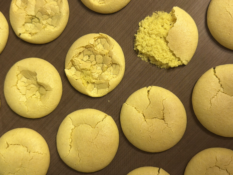 The image presents an overhead view of a collection of approximately twelve round, yellow baked goods, consistent with cookies or macarons, laid out on a dark reddish-brown silicone baking mat. Each item measures approximately 6-8 cm in diameter. Most of the cookies display various degrees of surface cracking. One cookie, situated in the bottom-right portion of the frame, is significantly fractured, exposing a vibrant yellow, crumbly, and porous interior structure. Another cookie, located near the center-right, exhibits a large indentation and extensive cracking on its upper surface. The remaining visible cookies show surface cracks. No individuals are present in the scene, and no specific actions are being performed. The environment appears to be an indoor setting, possibly a kitchen. The precise location within Tel-Aviv, Israel, is not discernible from the visual evidence.