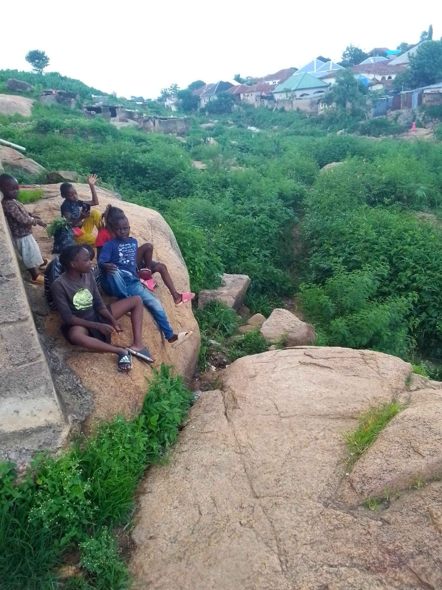 In Jos, Nigeria, a group of five children are gathered on a large, weathered rock formation. The rock is situated on a slope covered with lush green vegetation, and in the background, houses with corrugated iron and tiled roofs are visible on a hillside, indicating an urban or semi-urban settlement.
From left to right, a young child in a patterned brown shirt and white shorts stands near a concrete structure. Next to them, a child in a dark shirt and yellow skirt sits with their hand raised, appearing to wave. Another child, wearing a blue t-shirt and blue jeans, is seated on the rock, with their legs extended. To their right, a child in a dark t-shirt and black shorts is seated on the rock, looking to the left. Another child, partially visible, is also seated on the rock.
The children are casually dressed, and their poses suggest they are relaxing or interacting. The environment is natural with scattered rocks and dense greenery, interspersed with man-made structures in the distance. The lighting suggests it is daytime, possibly late afternoon, with a slightly overcast sky.