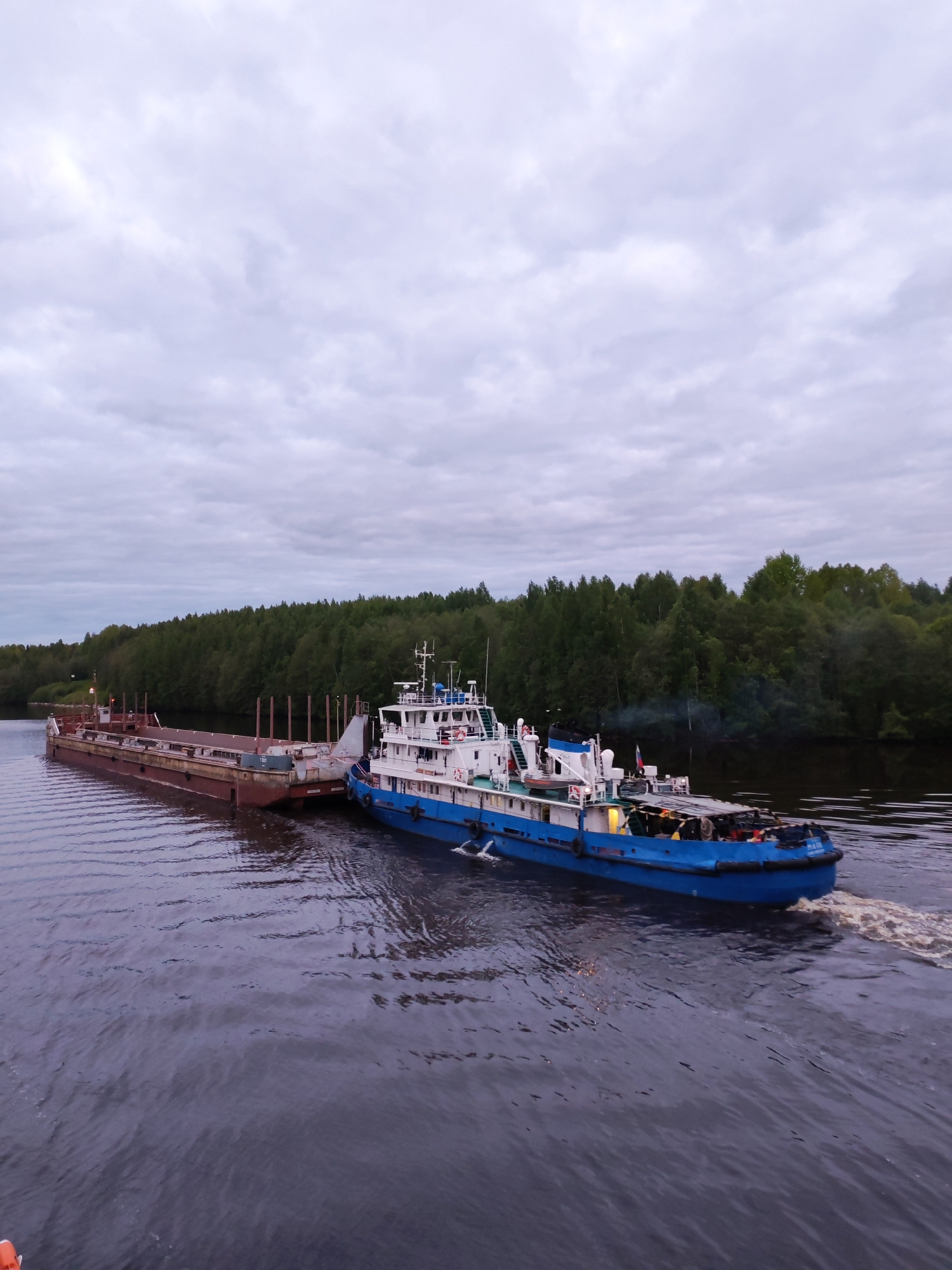 The image depicts a blue and white tugboat pulling a rust-colored barge on a river in Volokov Most, Russia. The sky is overcast and cloudy. The river is dark and has visible ripples and a wake trailing from the tugboat. Both banks of the river are lined with green trees, forming a dense forest in the background. The tugboat is attached to the barge at the front. There's some smoke coming from the vessel.