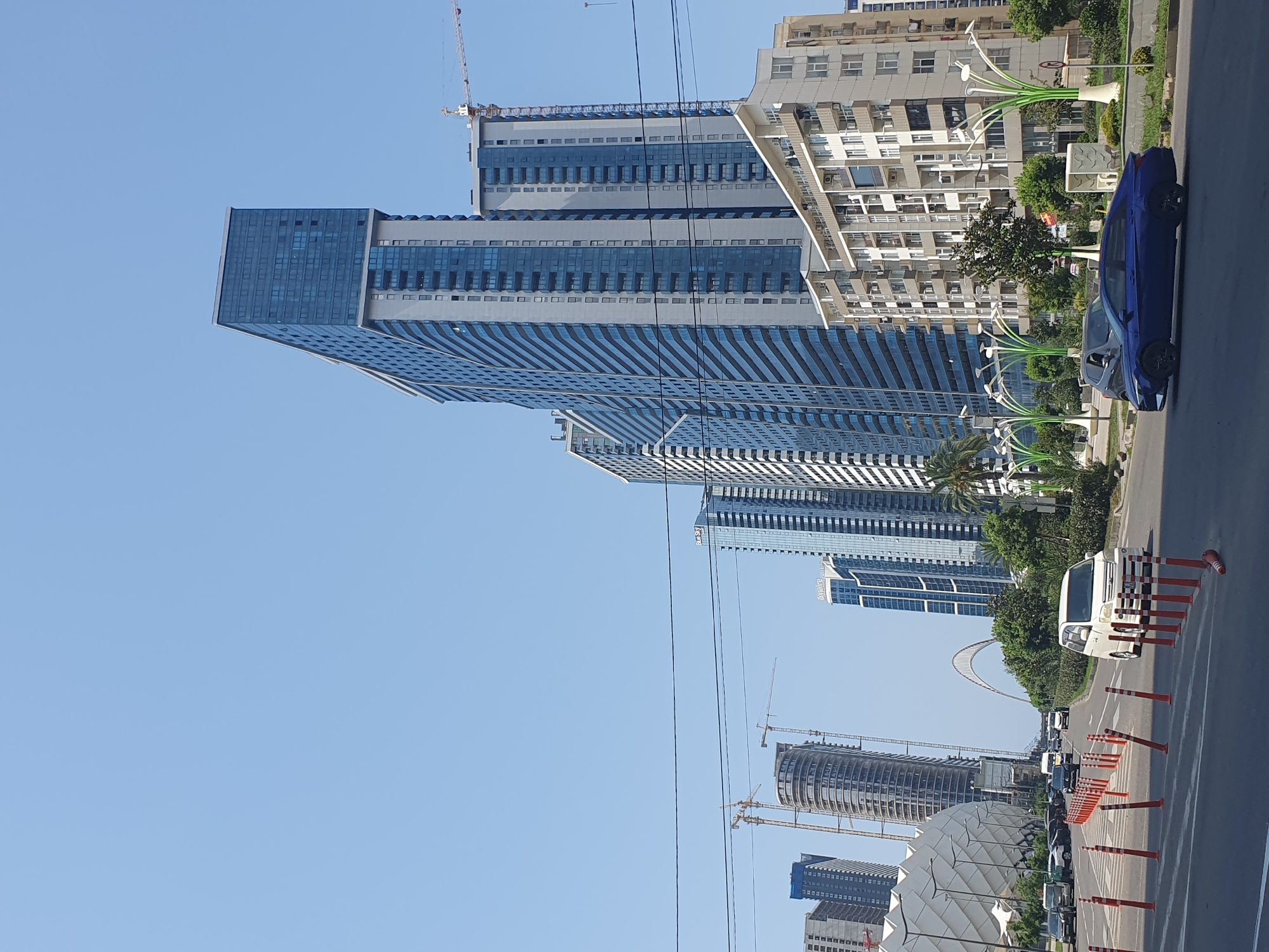This is a photograph of a street scene in Batumi, Georgia, featuring modern architecture and urban infrastructure. The image is dominated by a clear blue sky and a row of tall, contemporary buildings, many of which are clad in glass and feature balconies. The most prominent building on the right is a tall, slender skyscraper with a blue glass facade. To its left are several other high-rise buildings, some of which are still under construction, as indicated by the presence of construction cranes.
In the foreground, a multi-lane road is visible, with traffic cones delineating lanes and pedestrian crossings. Several vehicles are on the road, including a white minivan in the right lane and a blue sedan in the left lane. The road appears to be well-maintained, and there are pedestrian walkways alongside it.
Further in the distance, an arched bridge structure is visible, spanning across what appears to be a body of water or a wide avenue. The architecture on the left side of the image includes a large, white, curved structure with a distinctive patterned facade, possibly a stadium or an exhibition center.
The environment is characteristic of a coastal city undergoing significant development, with a blend of older and newer architectural styles, and ample green spaces, including trees and ornamental plants, lining the streets. The overall impression is one of a vibrant, growing urban center.