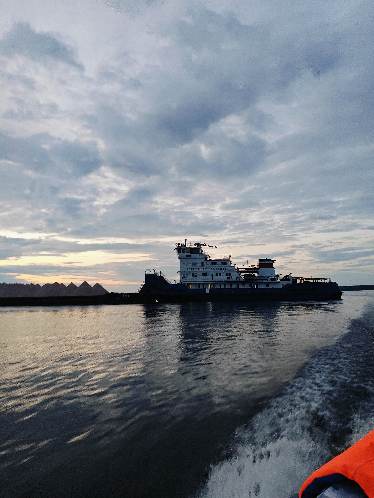 This image captures a wide shot of the Kama River in Chaikovsky, Russia, at dusk. A large tugboat, identified by the name "АЛЕКСАНДР РУМЯНЦЕВ" (Aleksandr Rumyantsev) visible on its side, is the primary subject. The tugboat is pushing a barge laden with dark, conical piles, likely of bulk material, such as sand or gravel. The river's surface is rippled, reflecting the overcast sky and the warm hues of the setting sun near the horizon. In the foreground, the wake of another vessel, possibly the one from which the photo was taken, is visible, with a splash of bright orange from a life vest in the lower right corner. The sky is filled with dramatic, layered clouds, typical of a twilight scene. The overall impression is one of industrial activity and transportation on the river during the transition from day to night.