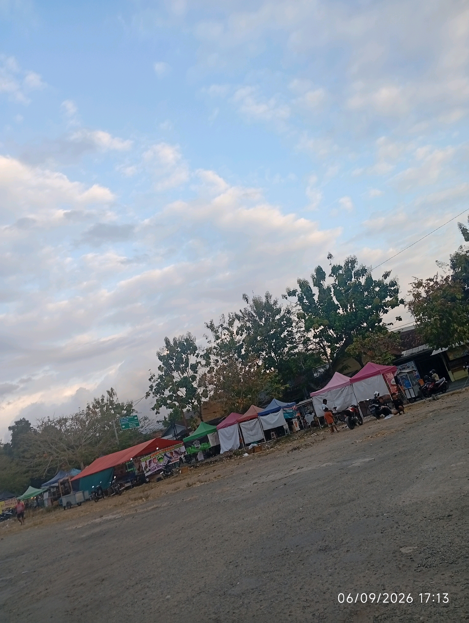 This is a medium-shot, outdoor, angled photograph of a street market scene in Purworejo, Indonesia, taken during daylight. The sky is a pale blue with scattered white clouds. In the foreground, a dusty, gravelly road slopes upward to the right. A line of market stalls with colorful awnings (red, green, pink, and white) stretches across the middle ground. Some of the stalls appear to be selling food, with one displaying a sign with Indonesian text. Several motorcycles are parked in front of or near the stalls, and a few people are visible, including one person walking on the left and a few individuals standing or sitting near the stalls. Trees with green foliage line the background, and a building with a tiled roof is visible behind some of the stalls. A green street sign is also present. A timestamp in the lower right corner reads "06/09/2026 17:13".