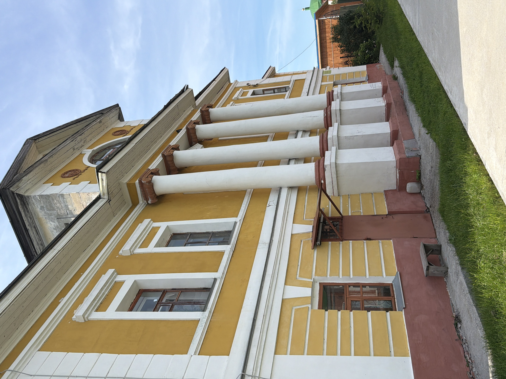 The image shows a side view of a yellow, multi-story building with white trim and a dark roof. The building has several windows with wooden frames and a series of large, white columns with red bases supporting an overhang. The lower portion of the building is painted a reddish-brown. To the left of the building, there is a small, wooden bench next to a doorway. The ground in front of the building is a concrete walkway with grass growing alongside it. In the background, there is a wooden fence and a building with a green dome. The sky is blue with some clouds. The architectural style suggests it is an older building, potentially a public institution or historical site. The location is identified as Usolye, Russia. There are no people visible in the image.