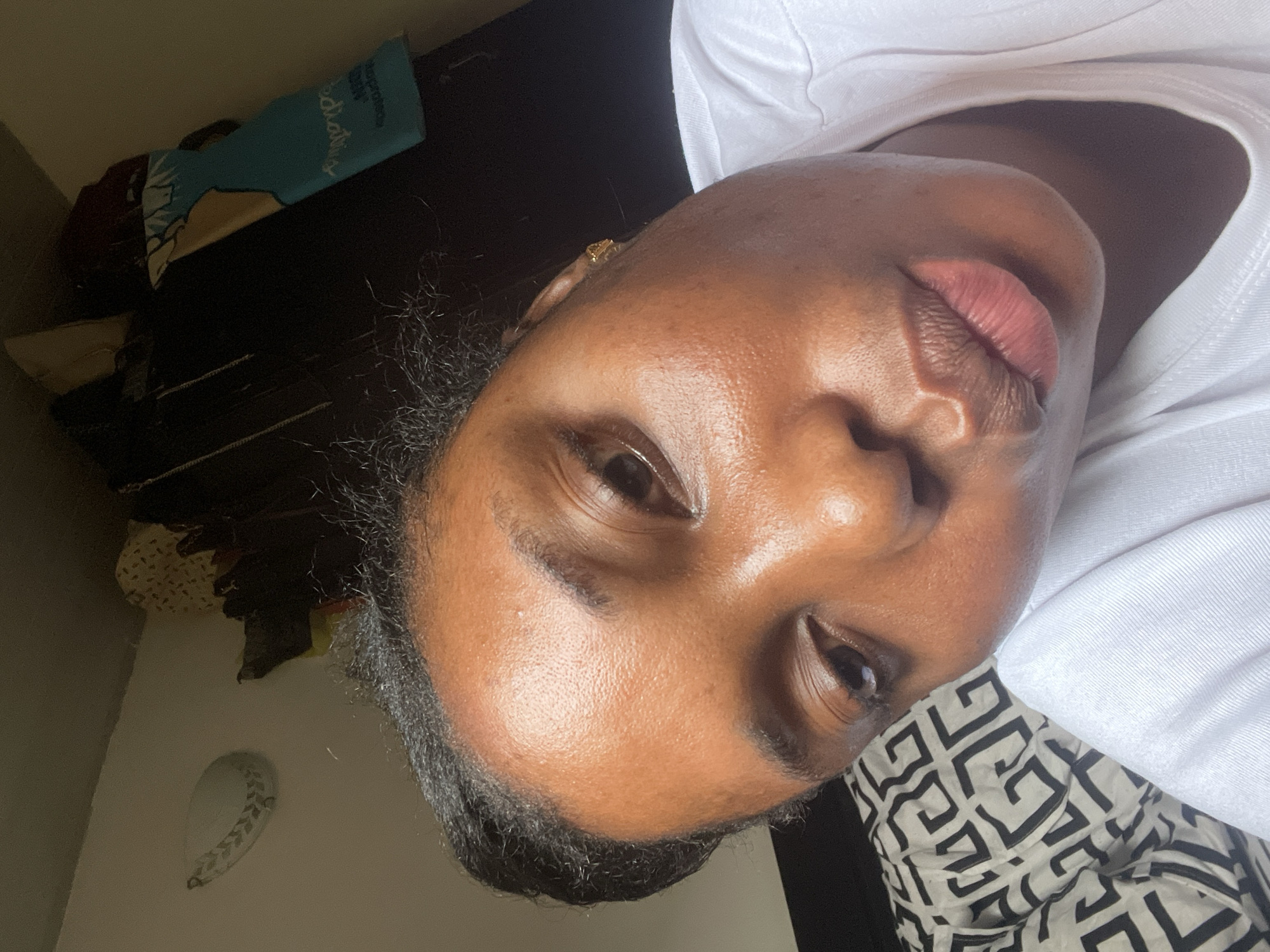 A close-up, rotated perspective shows the face of a dark-skinned woman. She has dark eyes and lips, a gold stud earring in her left ear, and is wearing a white t-shirt. The background is blurry, but includes dark furniture, a teal item, and a patterned textile. Given the prompt's request for a location in Abuja, Nigeria, this scene likely depicts the woman inside a residence or other indoor location within that city.