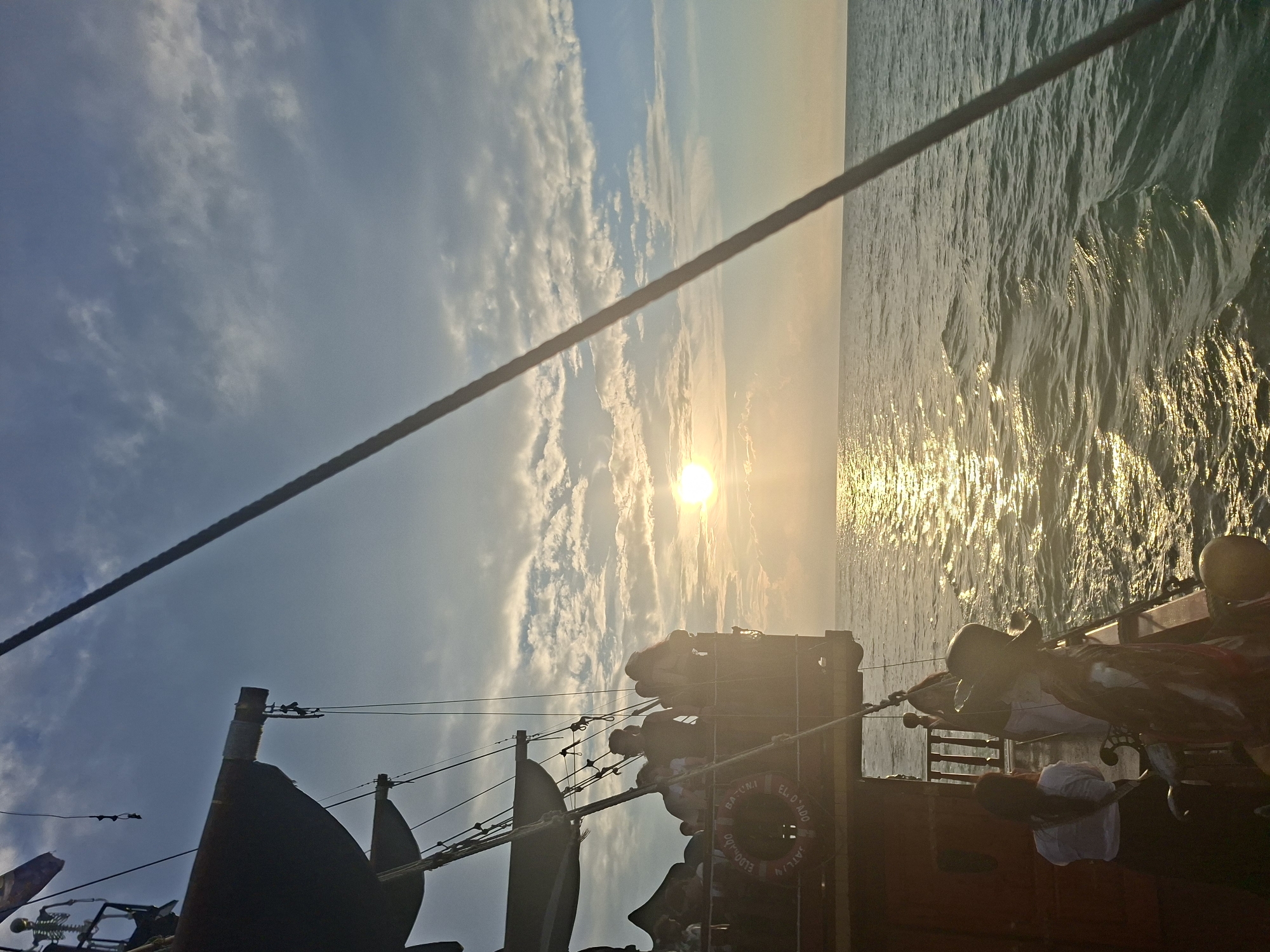 The image shows a view from a pirate-themed ship sailing on the Black Sea, with the sunset casting a golden glow on the water. The ship's wooden deck and railings are visible in the foreground, along with several people enjoying the ride. Some individuals are standing and looking out at the sea, while others are conversing. A life preserver ring with "EL DORADO BATUMI" inscribed on it is attached to the ship's railing, indicating the location of Batumi, Georgia. The sky is partly cloudy, with the sun visible on the horizon. The overall atmosphere suggests a leisurely excursion on a warm evening.