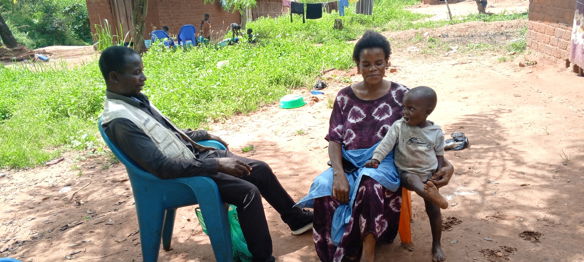 This image captures an outdoor scene in Makungu, Democratic Republic of the Congo, under bright daytime sunlight. In the foreground, a Black man sits in a blue plastic chair on the left, dressed in a dark shirt and trousers, with a light-colored vest over it that has faint text reading "AMW". He looks calmly to the right. To his right, a Black woman sits on a low orange stool or seat, wearing a purple and white patterned dress and a blue waist wrap. She holds a young Black child on her lap. The child, dressed in a light-colored long-sleeved shirt, has an animated expression with an open mouth and wide, curious eyes, looking towards the right.
The setting is a rural or village environment with a dirt ground interspersed with patches of green grass and vegetation. Distinct shadows indicate a sunny day. In the mid-ground, a green plastic basin rests on the dirt. The background features several other individuals, primarily children, scattered across a grassy area. Some are sitting on blue plastic chairs, while others are moving about, suggesting ongoing daily activities or play. A clothesline with drying clothes is visible near simple, possibly mud-brick structures or huts, adding to the depiction of everyday life in a communal outdoor space.