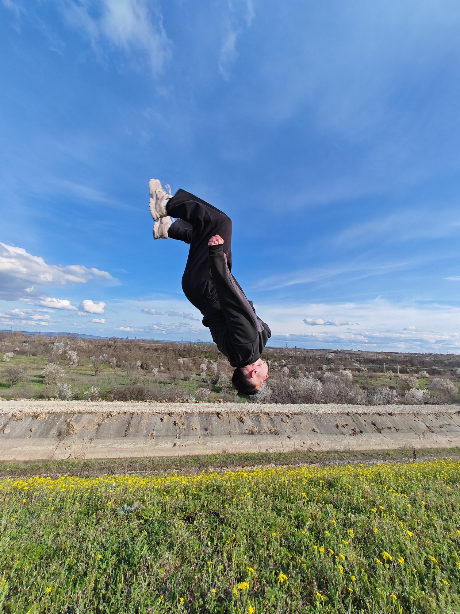 A person wearing black tracksuits and white sneakers is mid-air, performing a backflip. The sky is blue with scattered white clouds. In the background, there's a concrete structure running horizontally across the frame, likely a canal. Beyond that, there is a landscape of grass and trees, and blossoming fruit trees. The foreground is filled with green grass and yellow wildflowers.
This photo was taken near the city of Shumen in Bulgaria.