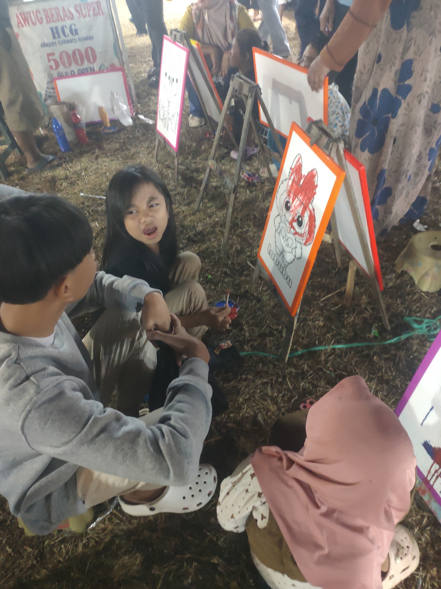 In Subang, Indonesia, at an outdoor event, two children are engaged in an art activity. A boy in a grey hoodie and white Crocs is seen from behind, his arm extended towards a girl. The girl, with long dark hair and wearing a black top and beige pants, is facing the boy with her mouth open, appearing to be speaking or reacting. They are seated on the ground amidst dry grass and fallen leaves, with several easels supporting white boards. One board displays a drawing of a red-headed cartoon character, and another has text that appears to read "labubu." Several bottles of colored liquid, likely paint, are scattered on the ground. In the background, other people are present, some standing and others seated, and a banner with text including "AWUG BERAS SUPER HCG" and "5000" is visible. The overall scene suggests a community or festival event involving children's art and entertainment.