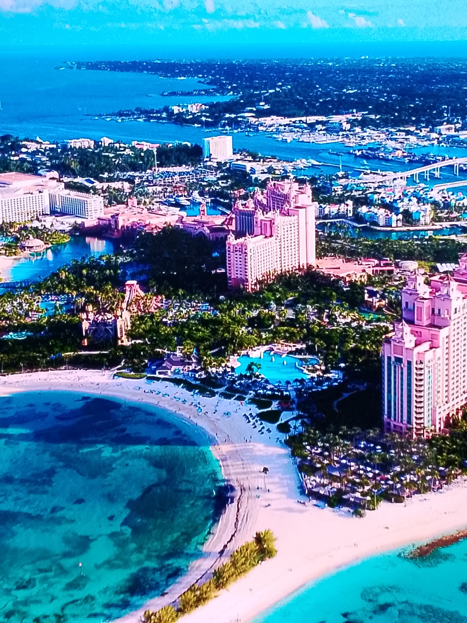 The image depicts an aerial view of a resort complex on a tropical island. In the foreground, a crescent-shaped sandy beach meets crystal-clear turquoise water. The beach is dotted with lounge chairs and umbrellas, and a few small figures are visible.
Beyond the beach lies a lush green landscape filled with palm trees, interspersed with buildings. The most prominent structures are several large, pink resort hotels with intricate architectural details. One hotel, in particular, features multiple towers and a central courtyard with a large pool. Adjacent to this resort, a complex of water slides and water attractions is visible, surrounded by greenery.
In the mid-ground, a harbor or marina is teeming with numerous boats of various sizes. A long bridge spans across the water, connecting different parts of the developed area. Further in the background, more buildings, including some white high-rise structures, are scattered amongst dense greenery. The horizon shows a vast expanse of the ocean, meeting a pale blue sky with a few scattered clouds.
There is no indication or visual evidence that this location is in Chișinău, Moldova. The environment, architecture, and vegetation are characteristic of a tropical coastal resort.
