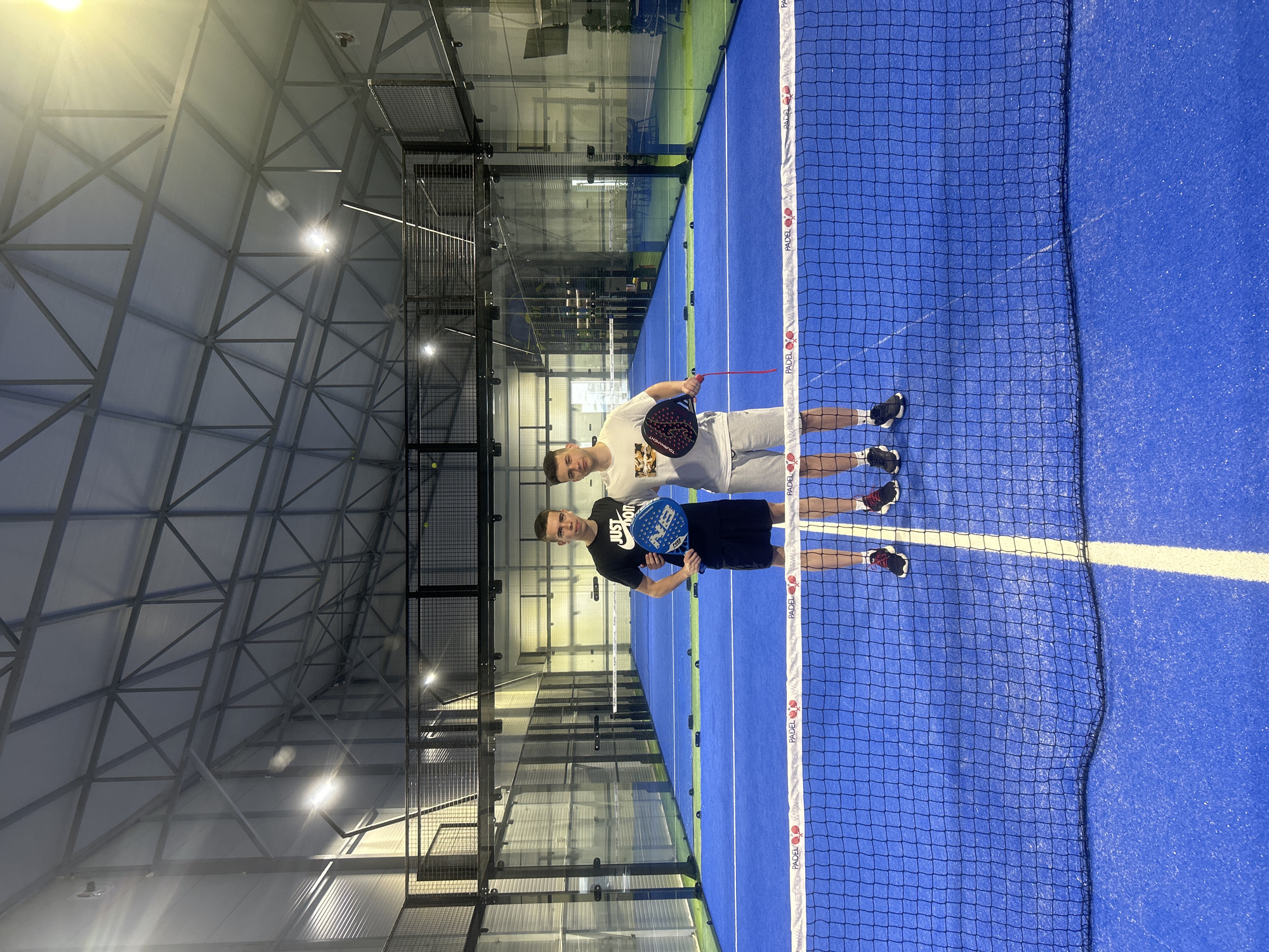 Two young men stand on a blue padel tennis court indoors, posing behind the net with their padel rackets. One wears a white t-shirt and light-colored shorts, and the other wears a black t-shirt and black shorts. The net is dark with white branding. The court is enclosed with glass walls. The building's ceiling is high and angular, with metal supports. The location is likely a padel tennis facility in Pleven, Bulgaria, based on the URL visible on the net.