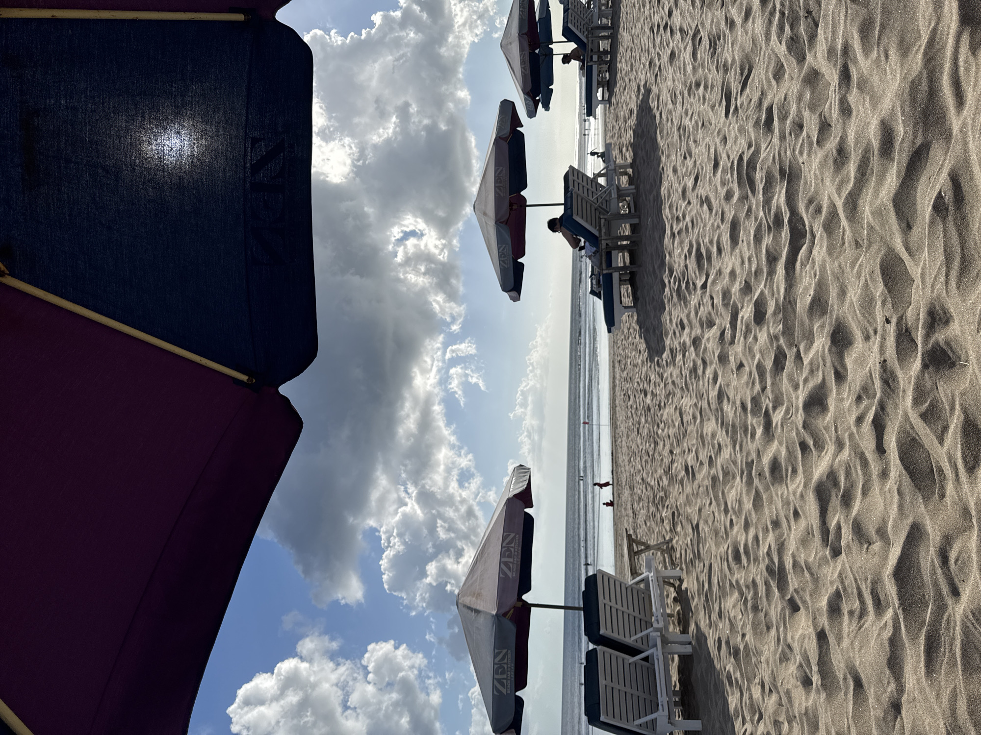 The image captures a scene at Kuta Beach, Indonesia, on a bright, cloudy day. In the foreground, the textured sand of the beach is visible, with shadows from unseen objects creating patterns. Two empty white lounge chairs are positioned under a gray umbrella with "ZEN BEACH BAR & KITCHEN" branding. Further back, more lounge chairs are occupied by people relaxing. One person is seated on a lounge chair under another umbrella, facing the ocean. In the distance, the sea stretches to the horizon, with small waves and a few figures wading or playing in the water. The sky is a vibrant blue, dotted with large, fluffy white clouds, some of which are illuminated by sunlight filtering through. The overall atmosphere is one of a relaxed beach day.