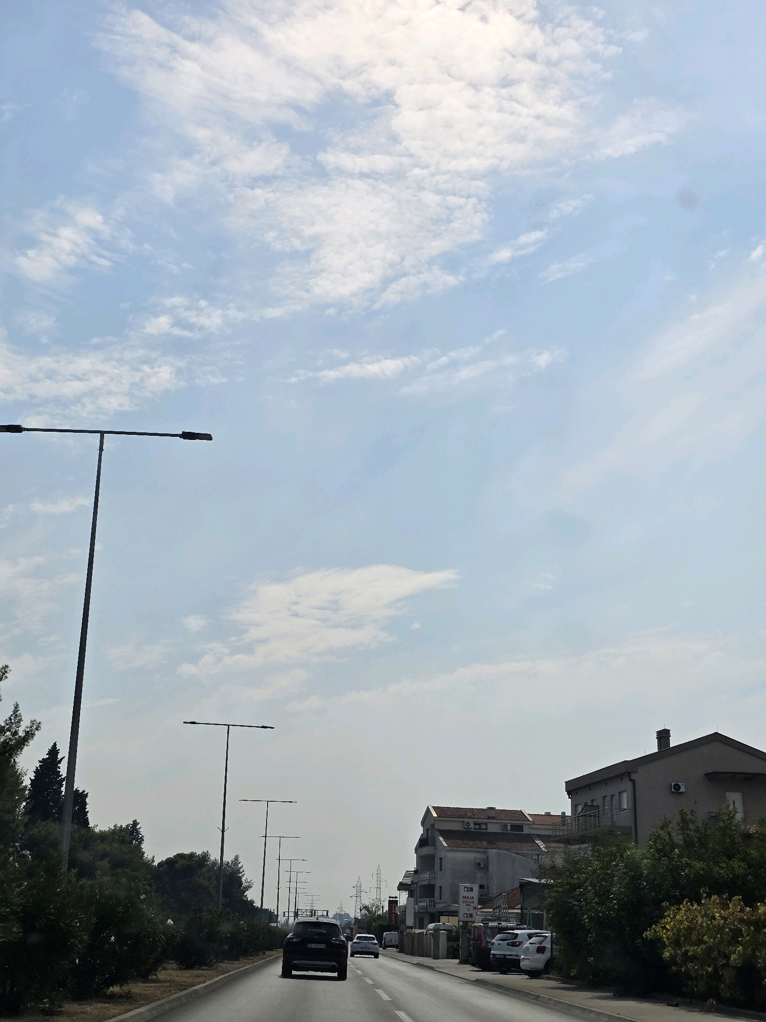 The image shows a street scene in Podgorica, Montenegro, under a partly cloudy sky. A road stretches into the distance, with several cars driving on it. To the left of the road, there is a row of trees and bushes, and beyond them, a tall street lamp. On the right side of the road, there are several buildings, some with balconies and air conditioning units visible. There are also more street lamps and power lines in the background. The overall impression is a typical suburban or semi-urban environment.