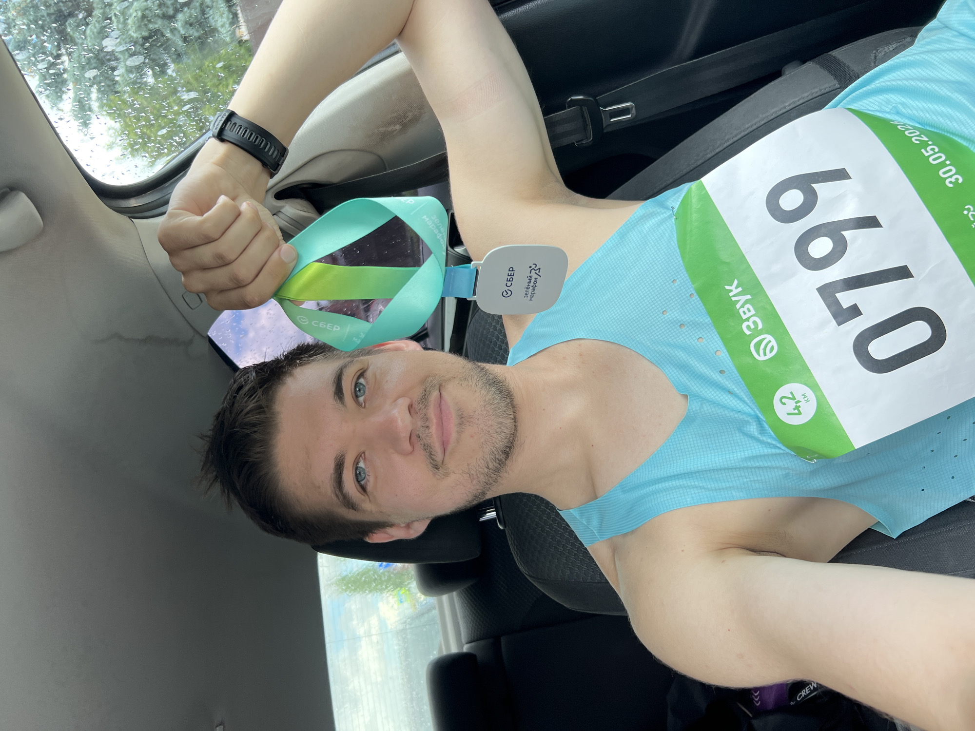 Here is a detailed description of the image:
A man with short brown hair and blue eyes is taking a selfie in what appears to be the passenger seat of a car. He is wearing a light blue athletic shirt and a black fitness tracker on his wrist. Attached to his shirt is a race bib with the number "0799" and the date "30.05.202". The bib also features logos for "Звук" (Zvuk) and Сбер (Sber). He is holding a green, blue, and white medal with the Сбер logo on the ribbon. The interior of the car is visible, with a dark gray seat, a seatbelt, and part of the dashboard. Through the car window, foliage can be seen, as well as rain on the glass. The medal also has the words "Зеленый марафон Сбер" (Sber Green Marathon) on it. The number of kilometers ran is also visible and it is 4.2 km. The race appears to be the Sber Green Marathon in Kazan, Russia.