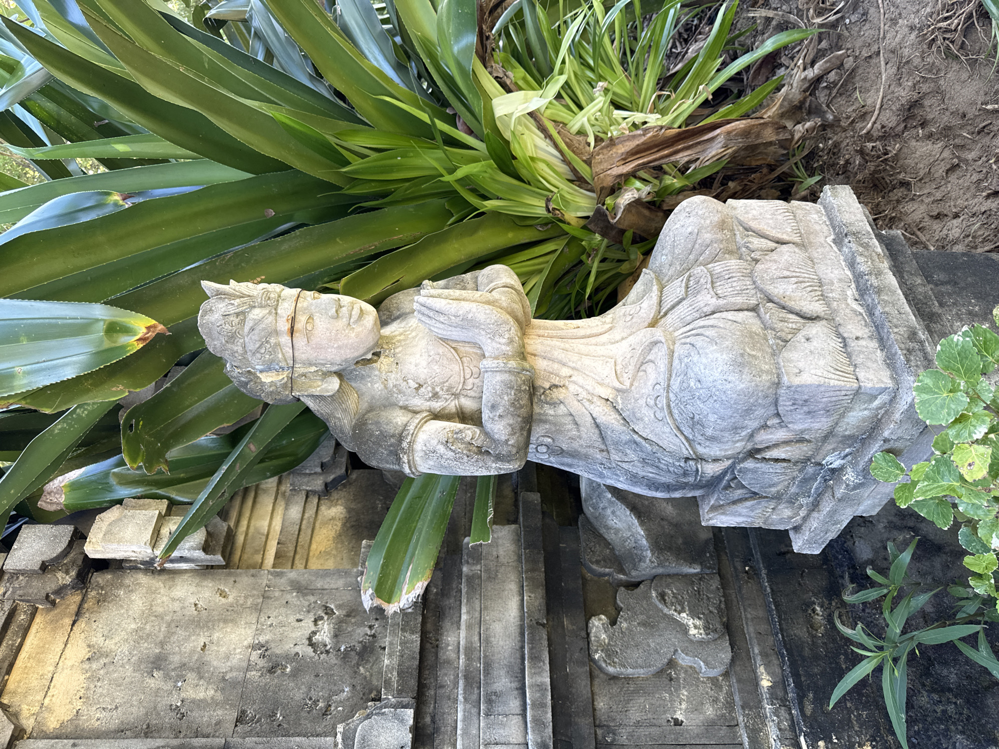 The image shows a stone statue of a person in a praying position, situated amongst greenery and stonework. The statue appears to be old, with visible weathering. It is positioned on a stone structure, possibly a wall or part of a building, which also shows signs of age and weathering. Various plants are seen around the statue and the stonework, including what looks like agave or aloe, and a few other leafy plants. The environment suggests an outdoor setting. Location is stated as Kuta Selatan, Indonesia.