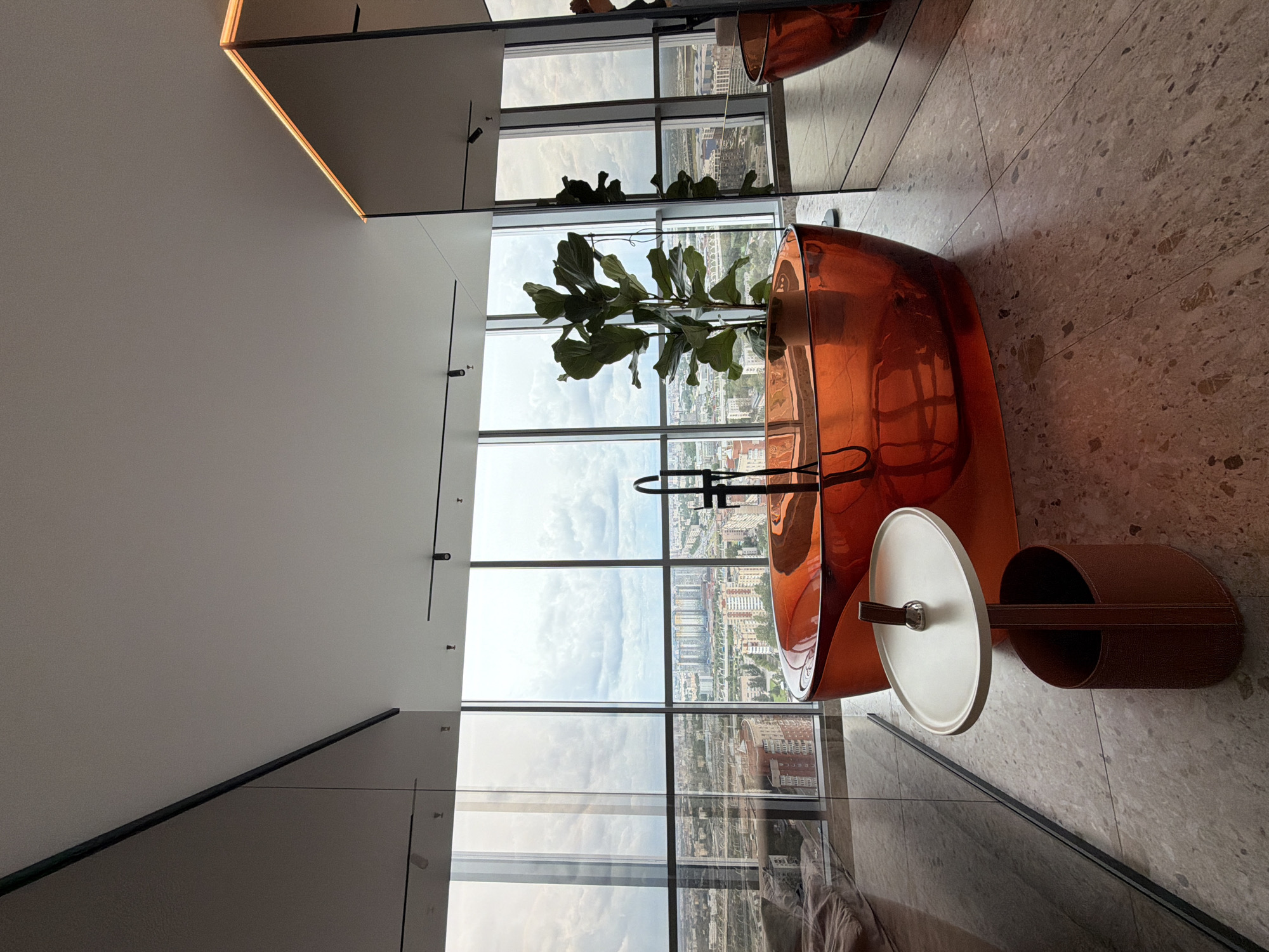 The image captures a luxurious bathroom with a panoramic view of Saint Petersburg, Russia. The room features an eye-catching, translucent orange bathtub, positioned to overlook a cityscape dominated by tall buildings and cloudy skies. Adjacent to the bathtub, a modern side table with a circular top and a brown, oval base stands on a speckled floor. A large potted plant with broad leaves adds a touch of greenery to the scene. The bathroom is illuminated by recessed lighting on the ceiling and accent lighting along a mirrored wall. Reflections in the mirrored surfaces reveal additional architectural elements and part of another orange bathtub. The overall impression is one of contemporary design and opulent living with a stunning urban backdrop.