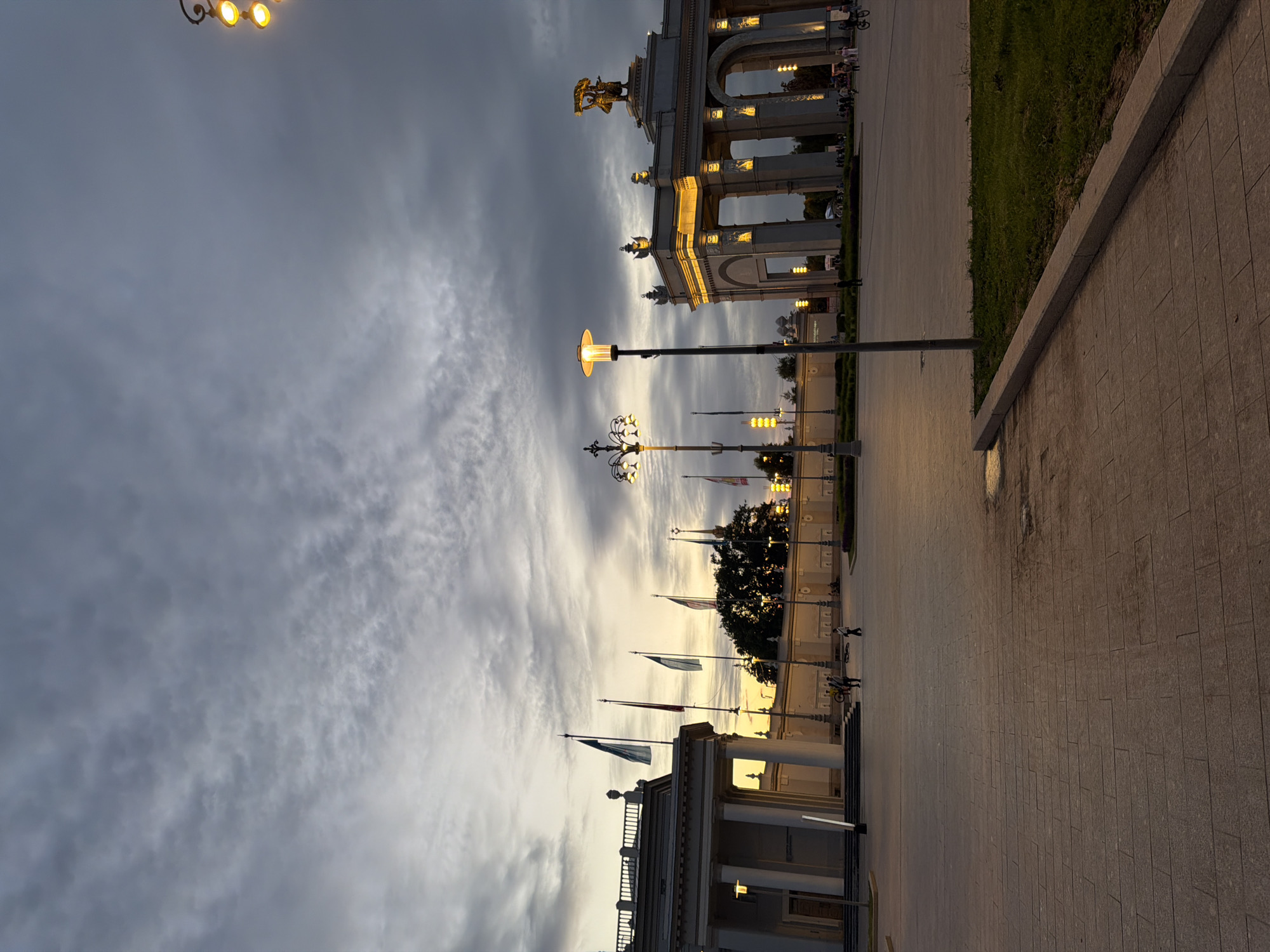 The image depicts an outdoor public space in Unknown, Unknown, likely at dusk or dawn, given the dramatic cloudy sky and the illuminated structures. In the foreground, a paved plaza stretches across the scene, with a strip of grass and a curb on the right side. Several people are visible in the mid-ground, appearing to walk or stand.
Dominating the left side of the mid-ground is a grand, white neoclassical building with a colonnade and a balustrade. Several flags are planted in front of this building, fluttering in the breeze. A tall, ornate lamppost stands in the foreground, casting a warm glow.
To the right of the mid-ground, a large, arched structure illuminated with yellow lights stands prominently. It is adorned with sculptures and decorative elements. In the background, a line of trees and more lampposts are visible. The sky is filled with heavy, grey clouds, with hints of yellow and orange suggesting the presence of the sun just below the horizon. The overall atmosphere is one of civic grandeur and subdued activity.