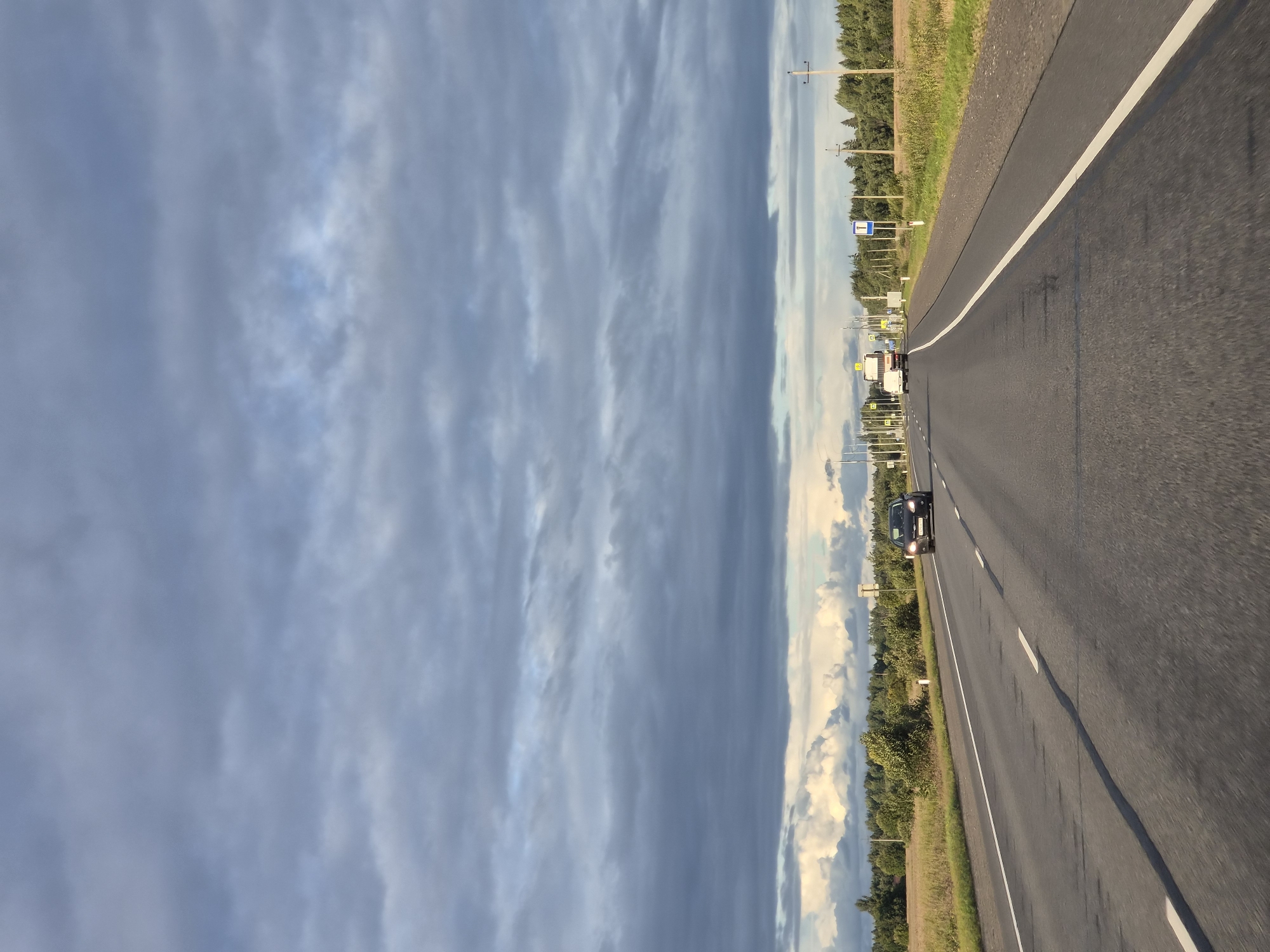 The image depicts a highway with a dark gray asphalt surface marked with white lane lines. The perspective is from within a vehicle, facing forward along the road. On the left side of the road, there is a field of dry grass. On the right side, there are trees and utility poles. The sky is filled with heavy, dark gray clouds, with some lighter, fluffy clouds visible in the distance near the horizon. There are several vehicles on the road: a dark-colored car is approaching in the right lane, and further ahead, a white truck is traveling in the left lane. Road signs are visible along the right side of the highway, including one with a blue square and a white "T" shape, indicating a turn. The scene appears to be in a rural area, possibly on the outskirts of Kipuya, Russia, given the presence of the highway, fields, and trees.