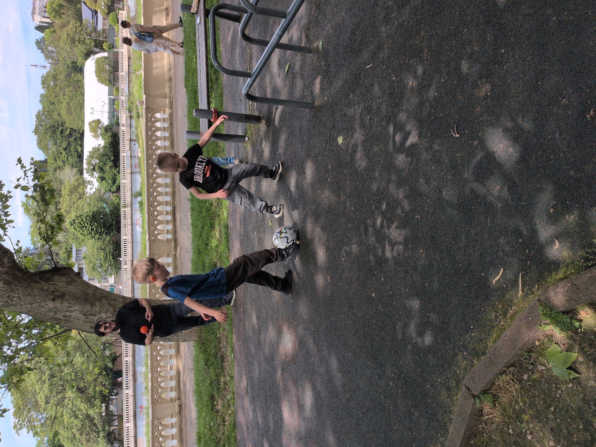 The image depicts a scene in a park in Sochi, Russia. Two boys are playing with a soccer ball on a paved surface near some playground equipment. One boy wears a blue shirt and dark pants and is in the act of kicking the ball. The other boy, wearing a "BROOKLYN" t-shirt and grey pants, stands nearby with his arms outstretched. An adult male stands near a tree to the left, holding an orange object. In the background, a decorative railing separates the park from a white building and treed area. Two people are visible in the background walking along a path.