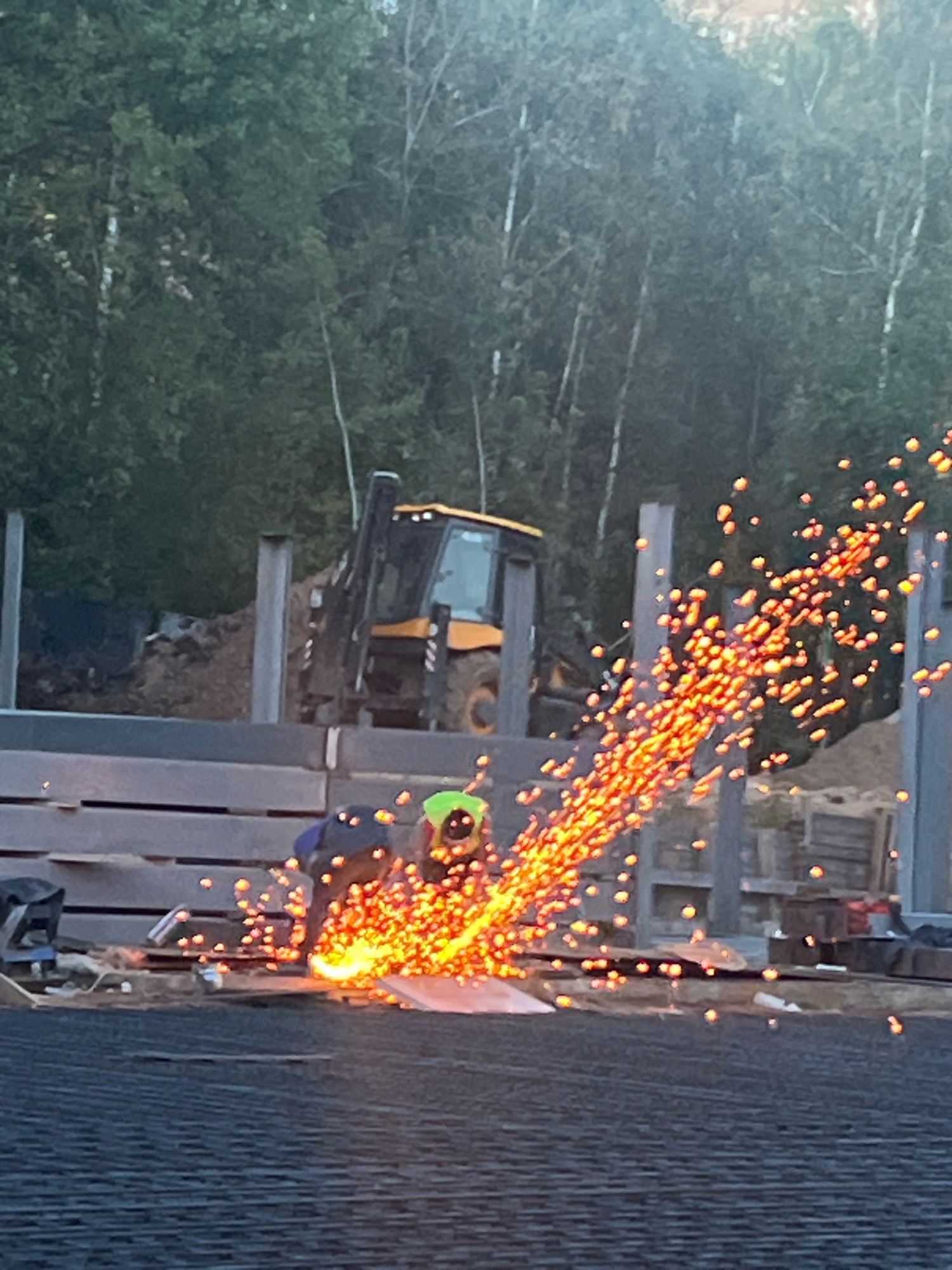 The image depicts a construction site with two individuals engaged in metalworking. One person, wearing a blue jacket and black pants, is operating a grinding tool that is emitting a large spray of sparks, illuminating the scene. Another person, wearing a neon green safety helmet and dark clothing, is also present nearby. In the background, a yellow backhoe loader is parked, and several large metal beams are erected, suggesting structural construction. The environment appears to be an outdoor setting with trees visible in the distance. There is no specific indication of the location being Zelenograd, Russia.