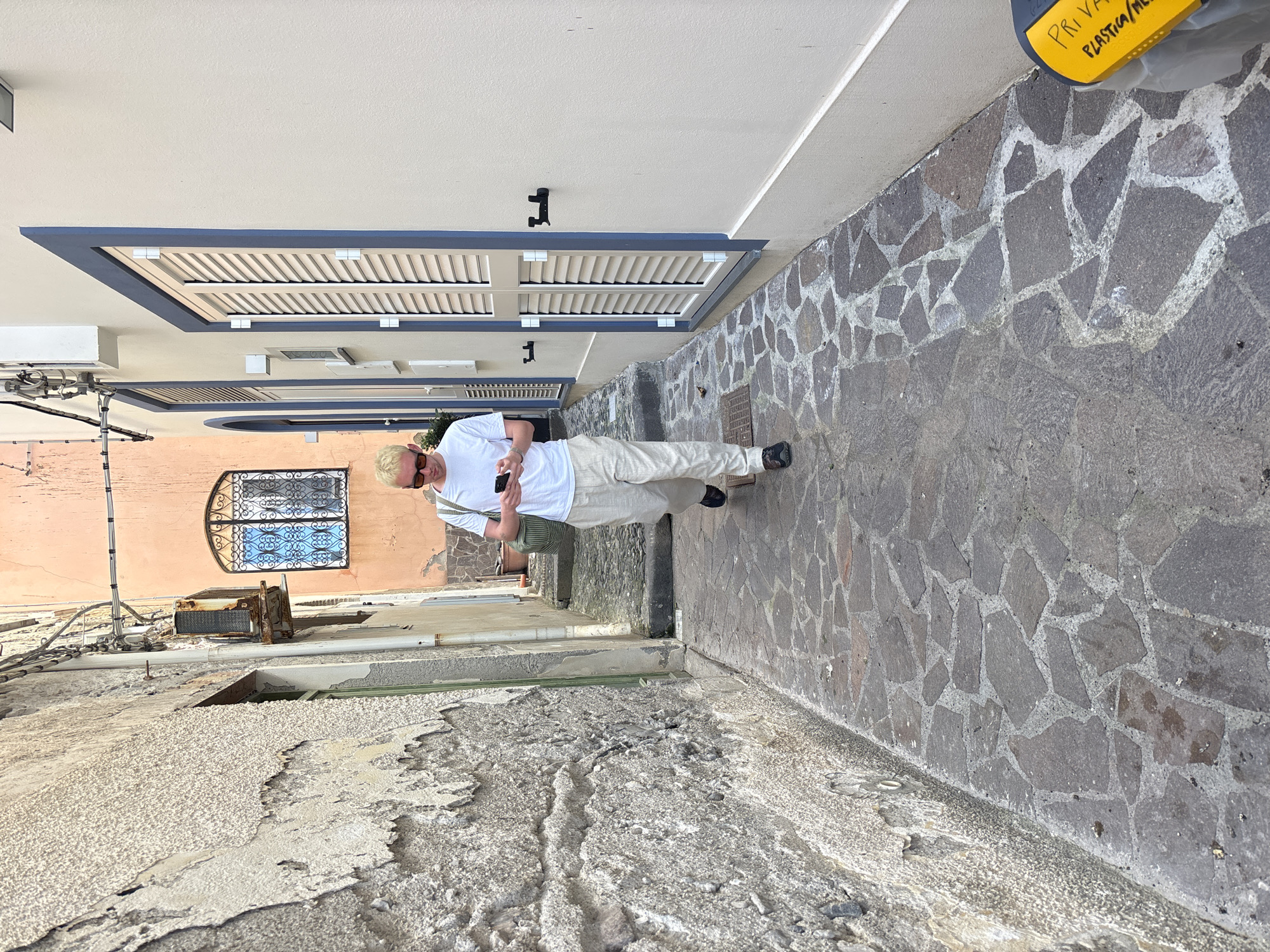 A man with blond hair and sunglasses, wearing a white t-shirt and light-colored pants, stands in a narrow, stone-paved street in Lipari, Italy, looking at his phone. The street is bordered by buildings; one has a white facade with blue-framed windows and another is peach-colored with an ornate window. The stone pavement is uneven and aged. The overall scene is bathed in bright daylight.