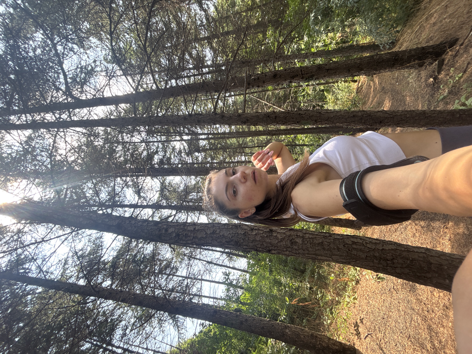 A young woman with brown hair pulled back in a ponytail takes a selfie in a forest. She is wearing a white crop top and dark leggings, and has a black arm band on her left arm. The forest is dense with tall pine trees, and the ground is covered in pine needles and dry grass. Sunlight streams through the trees, creating a dappled effect on the forest floor. The woman is looking directly at the camera with a neutral expression. The location is the pine forest of the Asparuhovo district in Varna, Bulgaria.