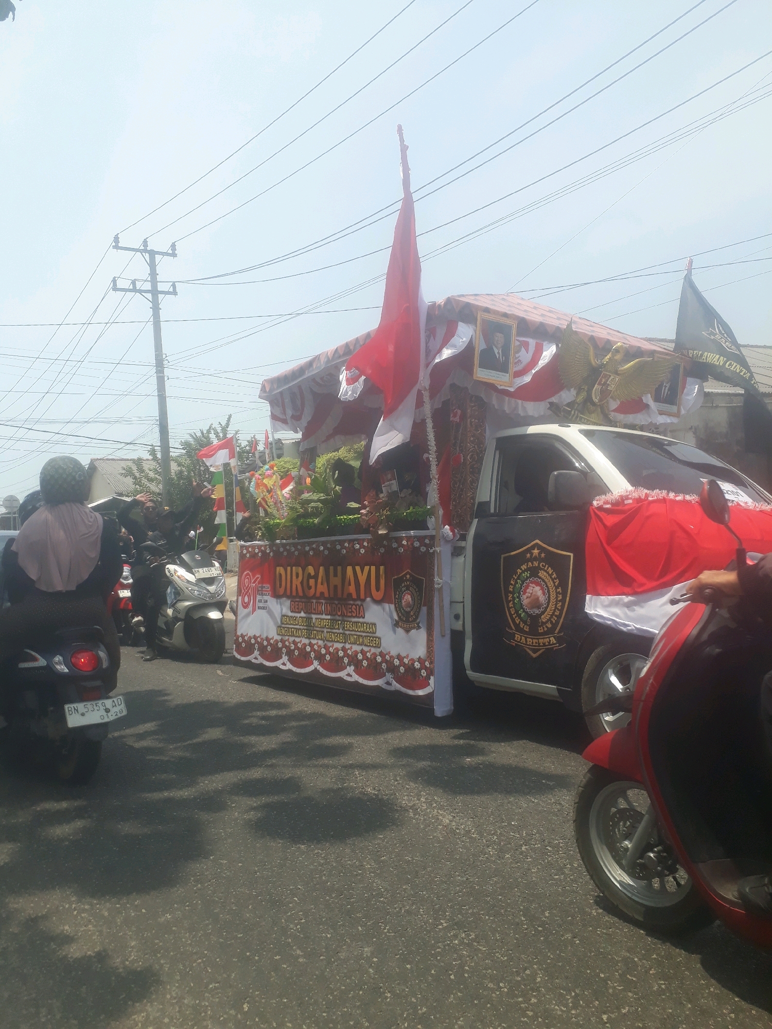 This is a medium-angle, eye-level, outdoor photograph taken on a sunny day, likely in Pangkalpinang, Indonesia. The image captures a parade or procession on a paved road, possibly during a national holiday celebration. The foreground features the rear of a dark-colored scooter with license plate "BN 5359 AD 01-28". To the right, a portion of a red scooter is visible, with its front wheel and leg shield in focus.
The main subject is a white pickup truck, heavily decorated for a parade. The truck is adorned with red and white Indonesian flags, a large banner proclaiming "DIRGAHAYU REPUBLIK INDONESIA" (Long Live the Republic of Indonesia), and floral decorations. Above the banner, a golden eagle emblem and framed portraits are visible. The truck also features a large shield emblem with the words "BARISAN CINTA TANAH" and "BARETTA".
Several people are visible on or near the truck, some waving. Other participants are on scooters, with one person in the foreground wearing a light purple headscarf and a dark helmet. Power lines and utility poles are prominent in the background, suggesting an urban or semi-urban setting. The overall atmosphere appears festive and celebratory.